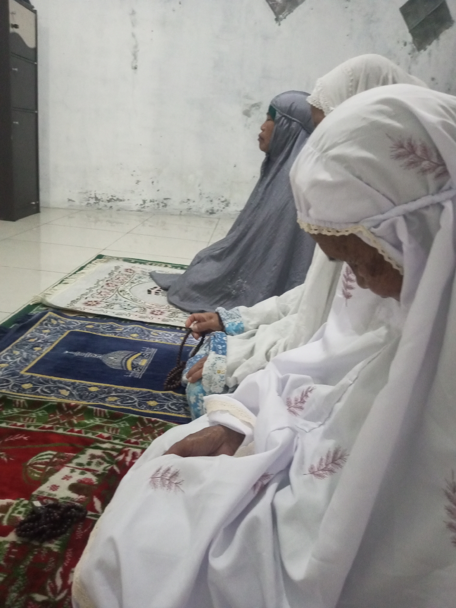Three individuals, dressed in Islamic attire, are engaged in prayer on prayer rugs placed on a tiled floor. The individuals are seated, with their heads bowed. One individual in the foreground is wearing a white hijab and a white garment with embroidered leaf patterns. They are holding a string of prayer beads. Another individual in the middle ground wears a grey hijab and a light-colored garment. The third individual, in the background, is wearing a white hijab. Prayer rugs with intricate patterns are visible on the floor. A tall, dark cabinet is positioned to the left of the scene. The wall behind the individuals appears to be plastered and shows signs of wear and tear. The lighting in the room is dim. This scene likely takes place in a prayer room or a home in Andongsili, Indonesia, given the context.