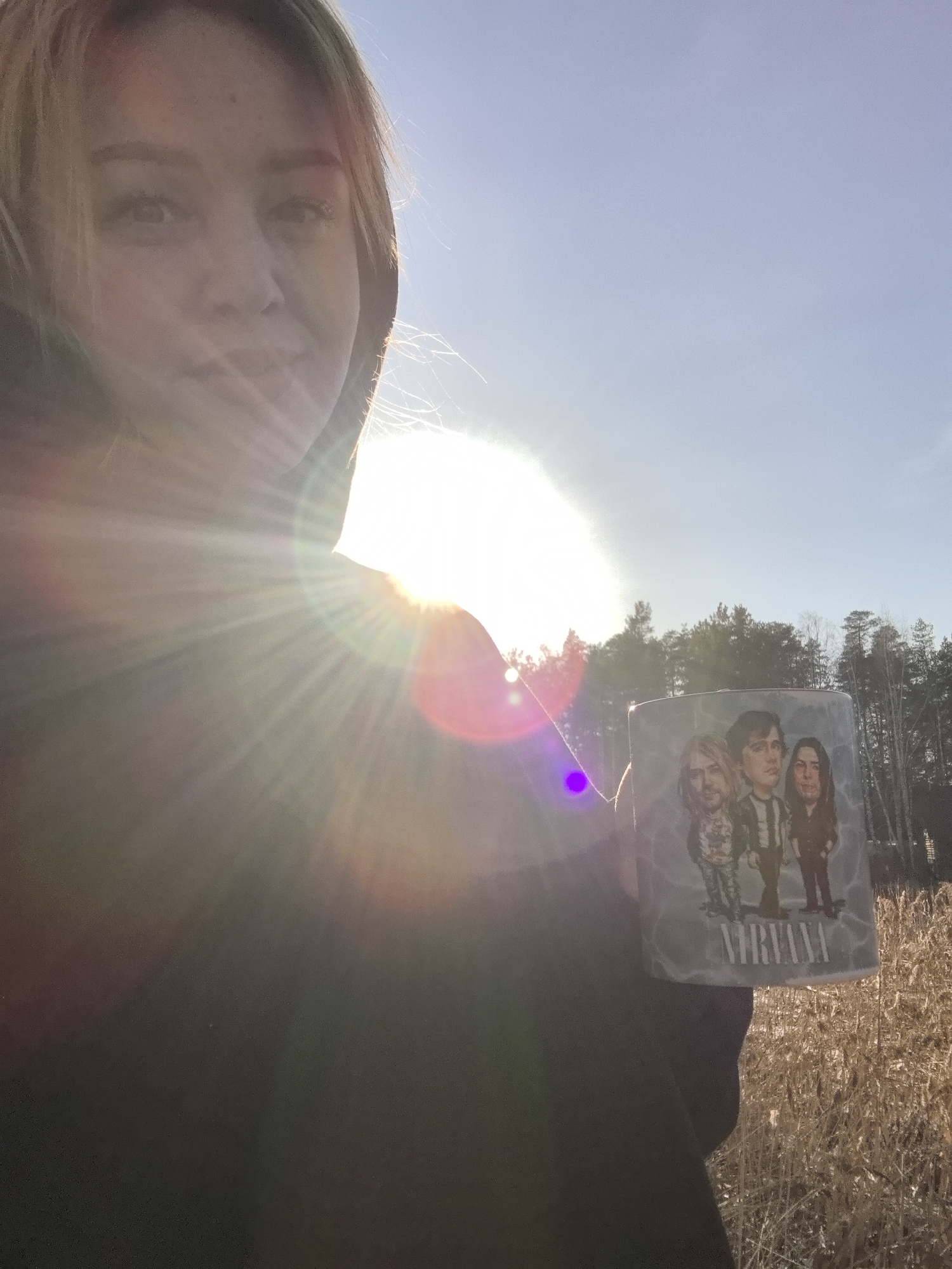 A blonde woman in a dark hoodie holds up a mug with a caricature of the band Nirvana on it, in a field of dry grass near a forest outside of Noviy, Russia. The sun is behind her, causing lens flare.