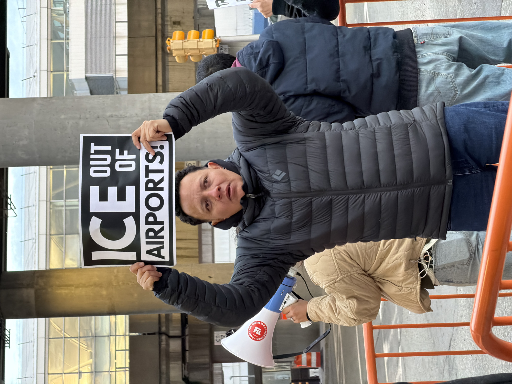 An outdoor scene captures a daytime protest under an elevated structure at John F. Kennedy International Airport (JFK) in New York City, United States of America. In the foreground, an orange metal barrier spans the bottom of the frame. Behind this barrier, a light-skinned adult male, with short dark hair, wearing a dark grey Black Diamond puffer jacket and dark blue jeans, stands facing forward, holding a white sign with black uppercase text that reads "ICE OUT OF AIRPORTS!" above his head. His mouth is slightly open, and he maintains a focused expression. To his left and slightly behind him, a partially visible individual in a beige quilted jacket holds a white and blue megaphone adorned with a red circular sticker that reads "PARTY FOR SOCIALISM & LIBERATION." To his right, another person is partially visible from the back, wearing a dark blue puffer jacket and light-wash Levi's jeans. In the background, a large concrete pillar supports an overhead elevated roadway or rail line. Further back, a multi-story airport terminal building with numerous glass windows and white horizontal paneling is visible. A vertical yellow traffic signal with three amber lenses is positioned to the right of the primary subject. Two orange and white striped construction cones are partially visible on the left side of the frame.