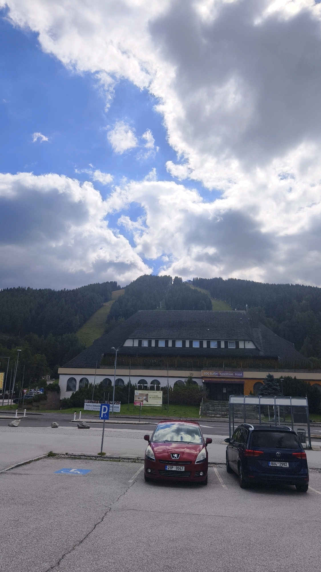 The image depicts a parking lot in front of a large building situated at the base of a tree-covered mountain with a ski slope visible. The sky is partly cloudy with patches of blue. In the foreground, two cars are parked: a red Peugeot 508 and a dark blue Volkswagen Touran. A blue handicapped parking sign is visible to the left of the red car. The building behind the cars appears to be a hotel, as indicated by signage that includes "BBQ Buffet. Konditorei Hotel". The mountain behind the building is densely forested, with a clear ski run descending from the summit. The overall scene suggests a resort or recreational area, likely in a mountainous region.
