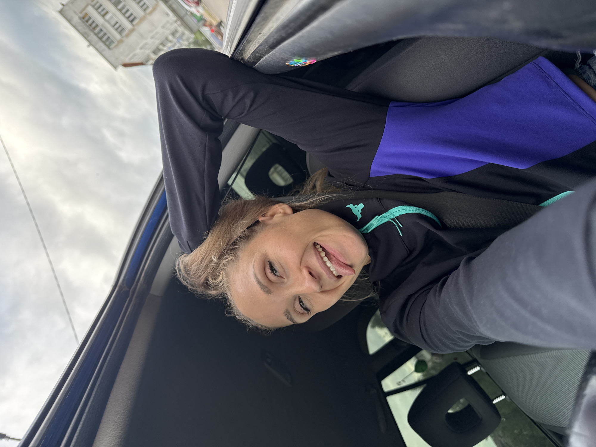 A young woman with blonde hair pulled into a ponytail is in the driver's seat of a car, sticking her tongue out at the camera. She is wearing a black and purple athletic jacket. Her left arm is resting on the open car door, and her right arm is behind her head. The background shows a cloudy sky and a multi-story building, possibly in Chaikovsky, Russia, indicated by the visible Cyrillic text on a building. The car interior is visible, including the passenger seat and headrest.