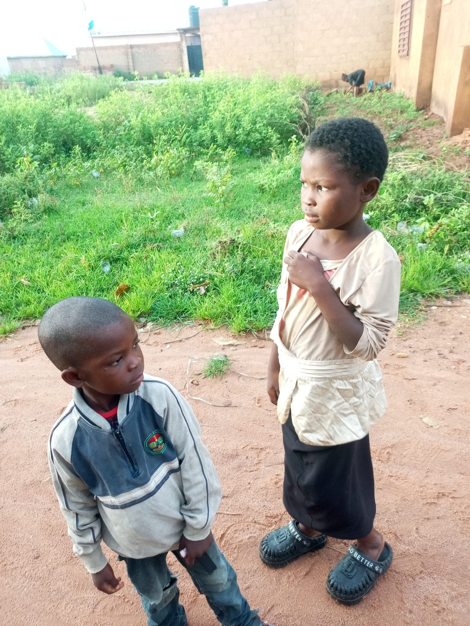 Two young children, a boy and a girl, stand on a dirt path in Tempure, Nigeria. The boy, on the left, wears a gray and blue zip-up jacket over a red shirt, and ripped blue jeans. He has a shaved head and is looking to his right. The girl, on the right, wears a tan shirt over a pink garment, a white wrap around her waist, and a black skirt. She has dark, curly hair and is looking to her left with her hand on her chest. In the background, there is a brick building with some greenery and more buildings in the distance.