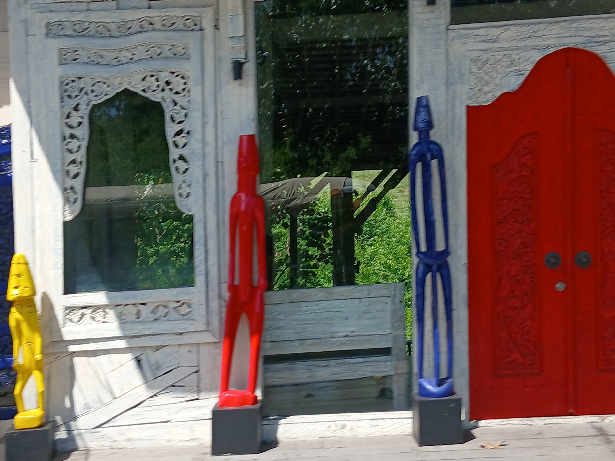 A bright yellow, humanoid wooden statue stands in front of a white ornate window frame. To its right, a red humanoid statue is positioned in front of a grey wooden bench. Further to the right, a blue humanoid statue stands beside a red double door. The scene is set in Mengwi, Indonesia, suggesting a cultural or artistic location.