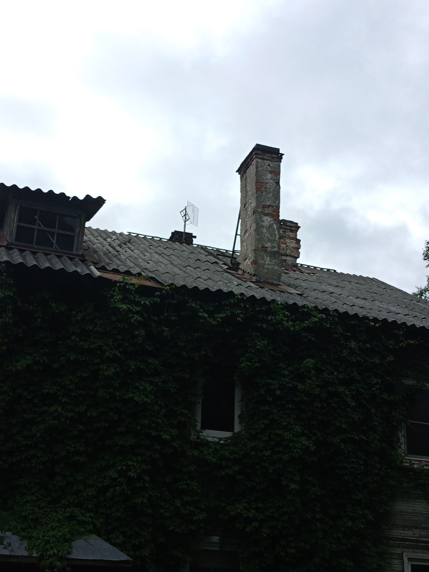 The image shows a close-up, low-angle shot of the upper portion of an old, weathered building, likely in a state of disrepair. The building is covered in dense green ivy, obscuring much of its facade. The roof is made of corrugated asphalt shingles, some of which appear damaged or missing. A dormer window with a broken pane is visible on the left side of the roof. A brick chimney, weathered and partially crumbling, stands prominently on the roof, with a metallic antenna attached to it. The sky is overcast and cloudy. The scene conveys a sense of neglect and the passage of time.