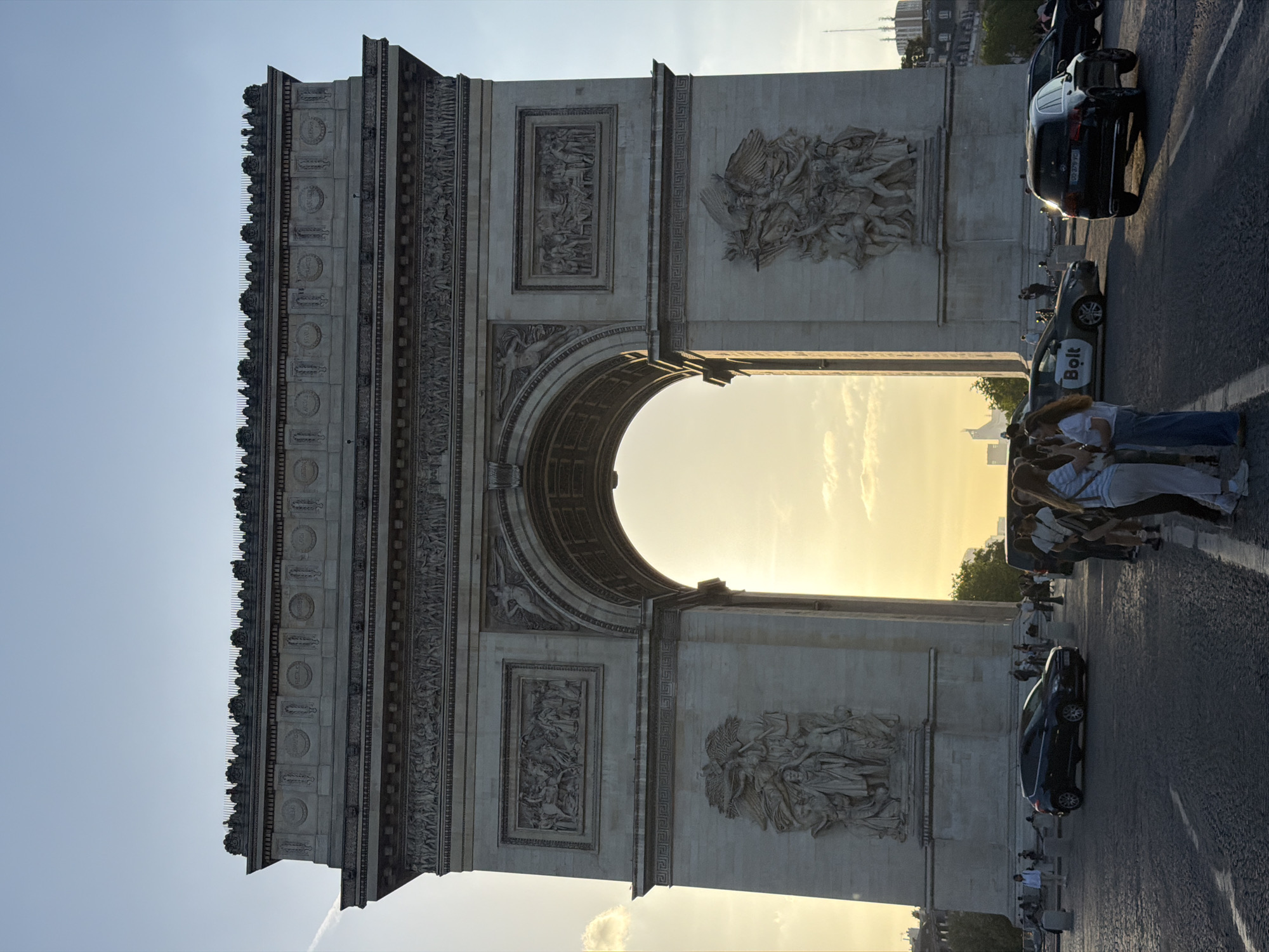 The image shows the Arc de Triomphe in Paris, France. The monument is made of gray stone and features detailed carvings and sculptures. People are on the right side, along with cars and taxis. The background contains trees and buildings, with a clear sky overhead. The light is bright, possibly indicating a sunset or sunrise.