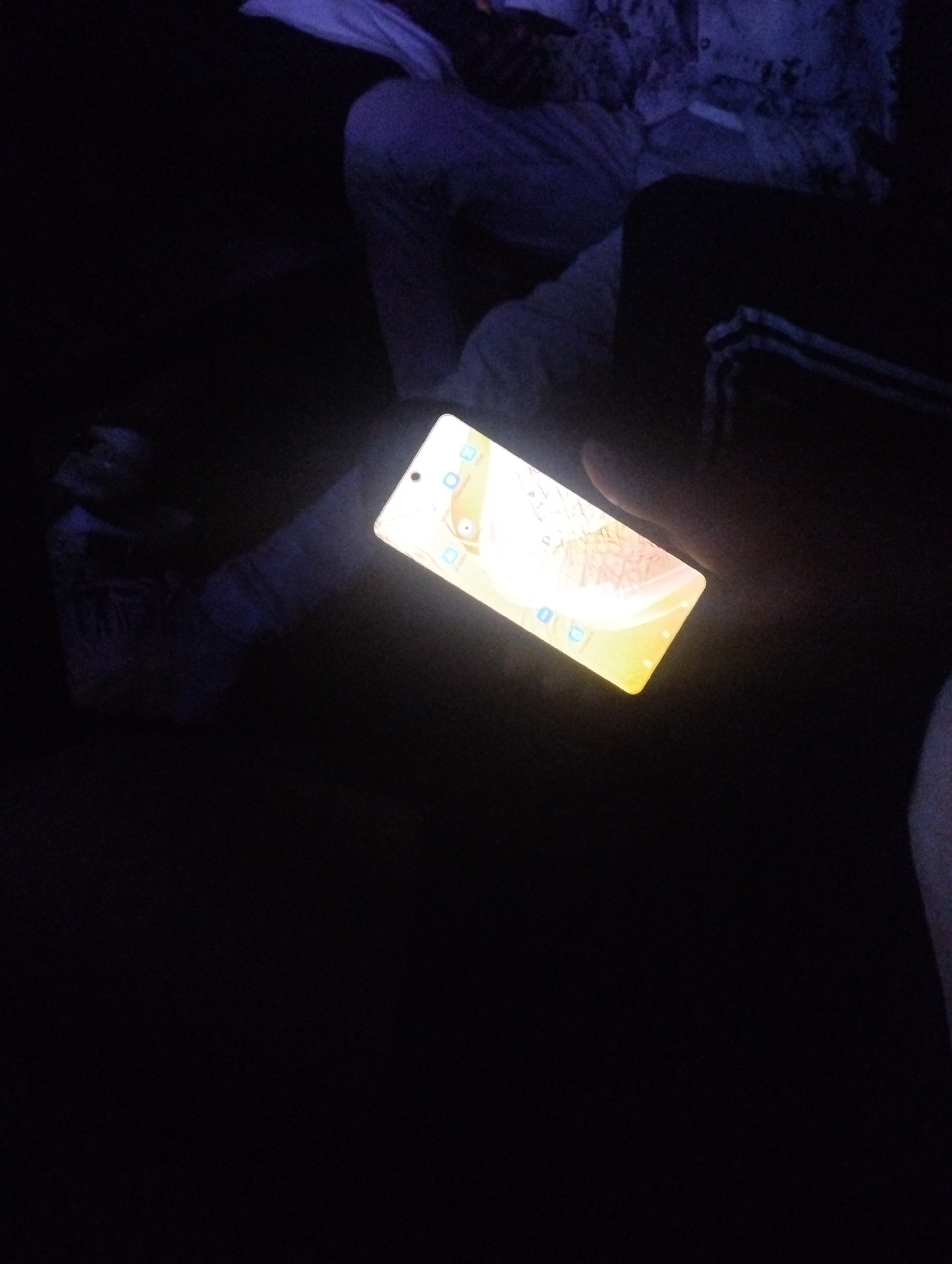 In a dark setting, a person is holding a brightly lit smartphone, possibly indoors. Only the phone's screen and part of the person's hands, legs, and clothing are visible due to the low light. The location is unknown, the city of Tumfure, Nigeria is not discernable in this image.