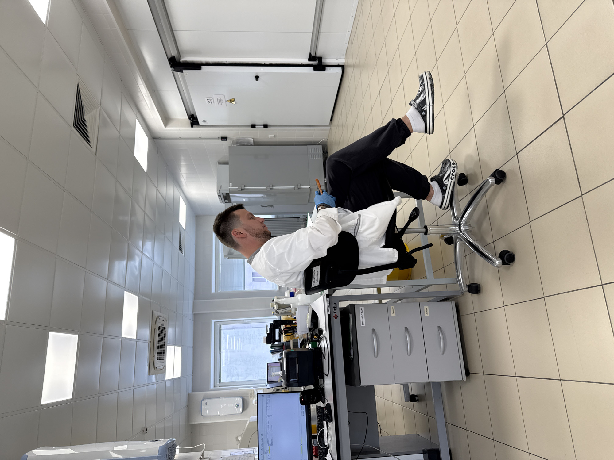 A person is sitting in a chair with their feet propped up in an indoor laboratory setting. The person is wearing dark-colored sweatpants, white socks, and black and white shoes. They are wearing a lab coat over their upper body and blue gloves on one hand. The room appears to be a clean, sterile environment with white walls and light-colored tile flooring. Laboratory equipment and computer monitors are visible on a table near the chair. There are cabinets, drawers, and a window in the background. The exact location in Moscow, Russia, cannot be determined with certainty.