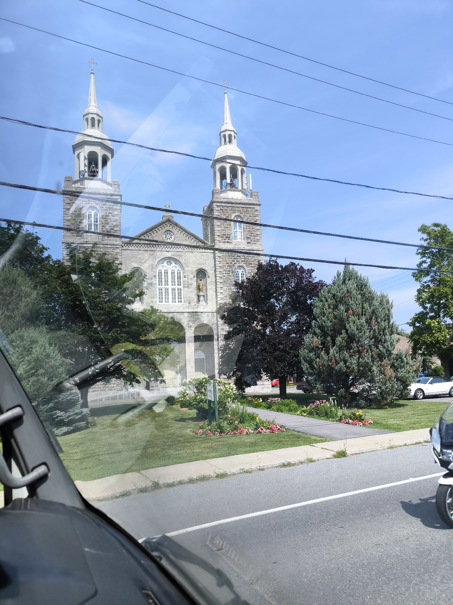 The image depicts a stone church with two bell towers, each topped with a spire and a cross. The church has a prominent arched entrance and several arched windows. The building is surrounded by a lawn with various trees and flower beds. A white convertible car is parked on the right side of the image, and a portion of a motorcycle is visible in the foreground. The scene appears to be taken from inside a vehicle, as indicated by the presence of a windshield wiper and the reflection of overhead wires on the glass. The sky is blue with some scattered clouds. The provided metadata indicates the location is Sainte-Martine, Canada.