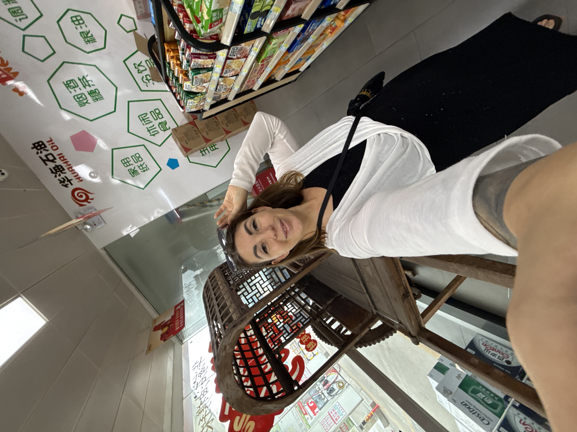 A woman takes a selfie in a convenience store, possibly at a gas station in Huizhou, China. She is wearing a black dress and a white cardigan, with sunglasses perched on her head. In the background, shelves are stocked with various products. A decorative wooden cart with red lanterns is also visible. The store's wall features hexagonal icons with Chinese text, one of which reads "HUA HAI OIL".