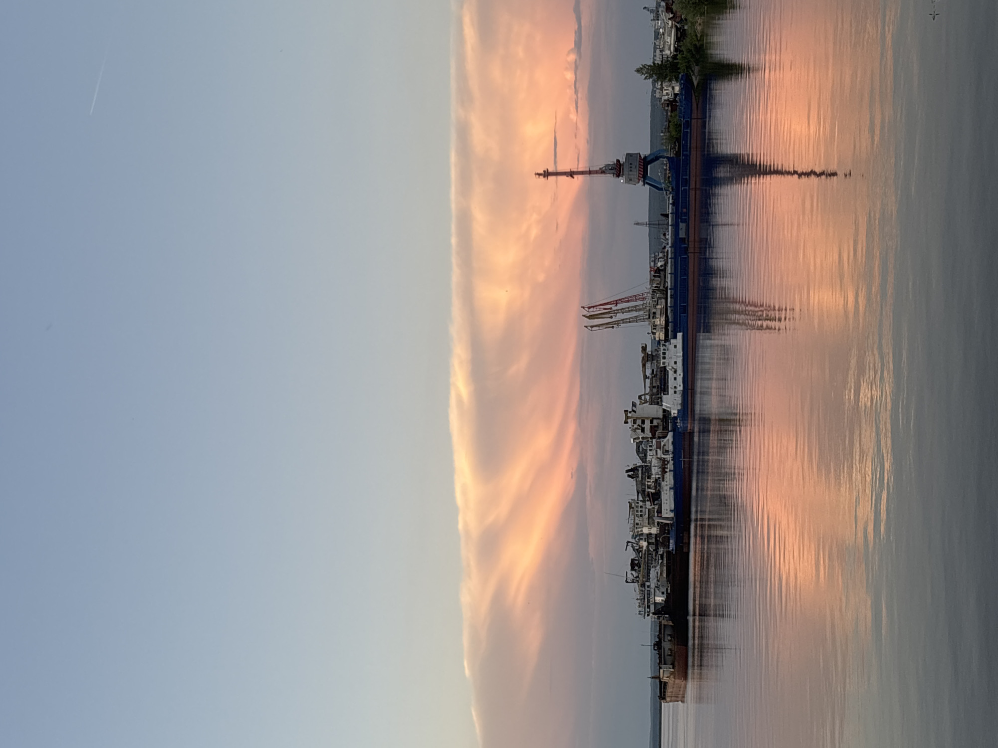 A panoramic, vertical, eye-level shot captures a sunset over a body of water, likely the Kama River, in Chaikovsky, Russia. The sky is a gradient of soft blues fading into a vibrant pink and orange as the sun sets behind a long, striated cloud formation. The water's surface is calm, reflecting the colorful sky and the silhouettes of docked industrial vessels.
Dominating the scene is a fleet of barges and ships moored along the shore. The vessels are a mix of sizes and purposes, with some appearing to be cargo ships and others possibly industrial or construction-related. Among them, a prominent blue and red port crane stands tall, its structure clearly defined against the twilight sky. The reflective surface of the water mirrors the crane and the warm hues of the sunset, creating a symmetrical visual effect.
In the distance, a faint contrail from an aircraft is visible in the upper right quadrant of the sky, indicating activity above. The far bank of the river is lined with trees and appears to be a mix of natural landscape and possibly more industrial structures, though they are indistinct due to the distance and lighting conditions. The overall impression is one of industrial activity harmonizing with the natural beauty of a sunset. No people are visible in the frame.