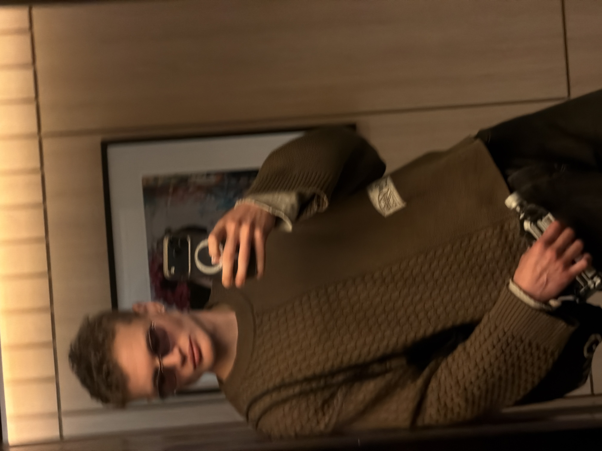 A young man with short, curly brown hair and sunglasses is taking a selfie in a mirror. He is wearing a brown textured sweater with a white tag. He is holding a black smartphone in his left hand and a bottle in his right hand. A framed picture hangs on the wall behind him. The location is possibly in London, United Kingdom, but this is not definitively clear from the image.