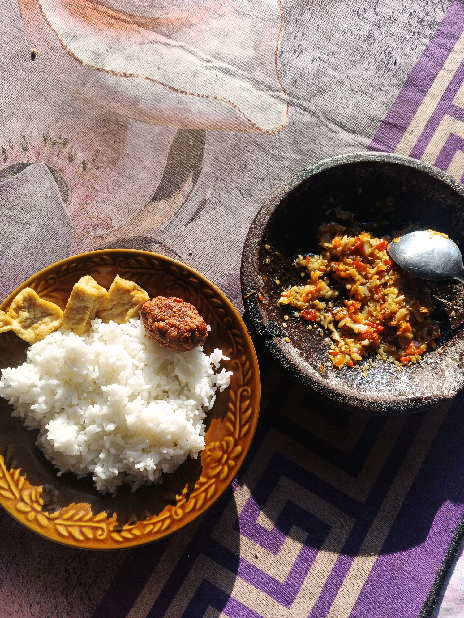 A top-down, eye-level shot captures a meal laid out on a patterned cloth. To the left, a brown ceramic plate overflows with fluffy white rice, topped with three pieces of golden-brown fried tofu and a single, round, dark brown fried patty. To the right, a dark, rough stone mortar holds a vibrant red and orange chili paste, with a silver spoon resting inside, ready to serve. The background features a textured cloth with abstract floral patterns on the left and a geometric design of purple and beige stripes on the right. The scene is illuminated by natural light, casting distinct shadows across the cloth, suggesting it is daytime. The setting is described as being in Bocek, Indonesia.