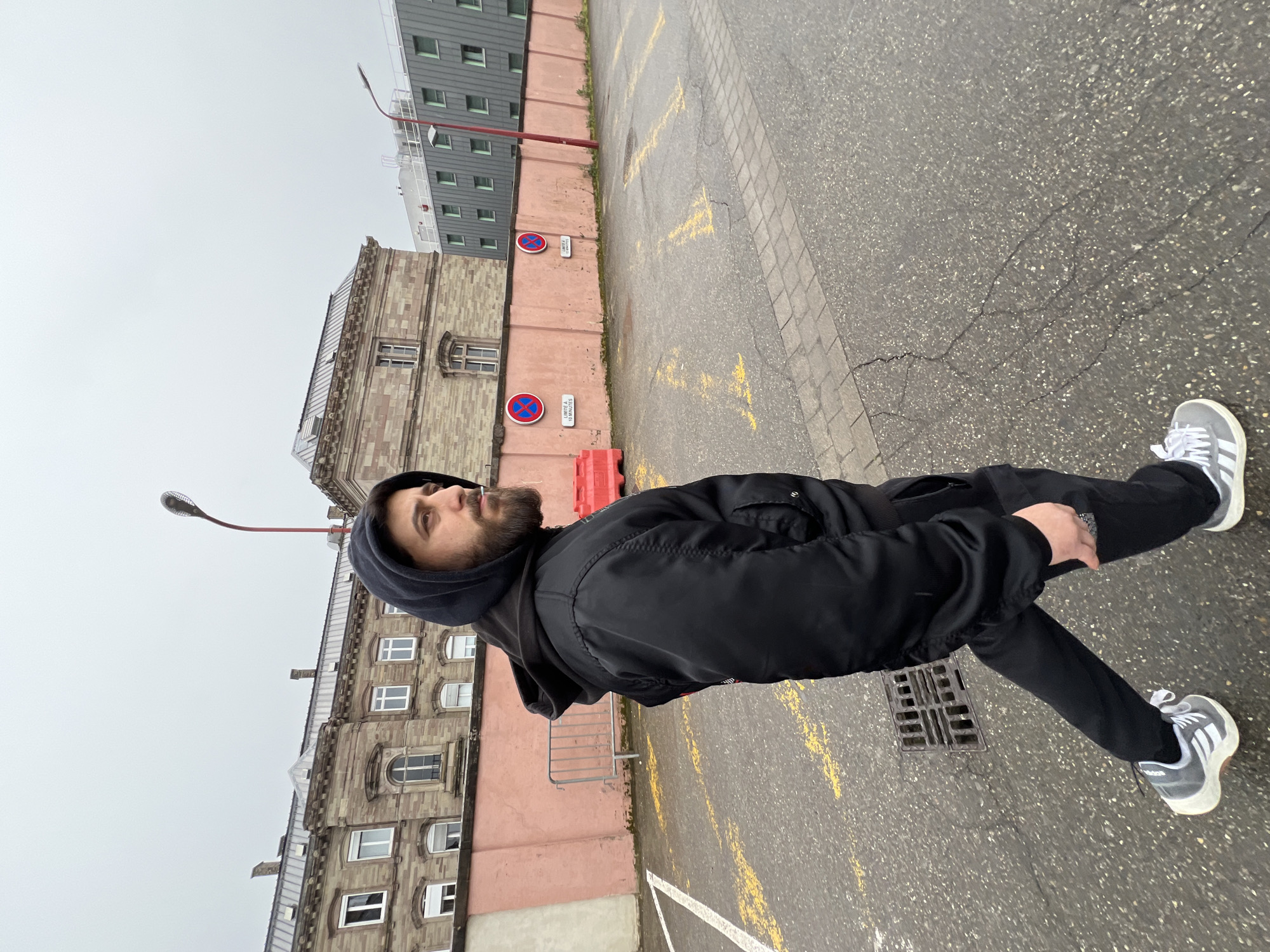 A man wearing a black hooded jacket, black pants, and grey Adidas sneakers is standing in a parking lot in Strasbourg, France. The lot is asphalt with yellow parking lines and some cracking. He is standing next to a storm drain. Behind him is a building constructed of brick and stone with several windows. Part of a more modern, grey building can be seen above the brick building. There are street signs prohibiting parking attached to the building. The sky is overcast.