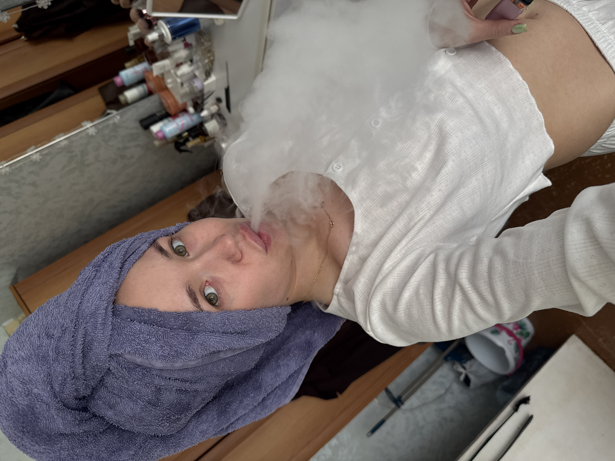 An indoor scene in the city of Chaikovsky, Russia, depicts a light-skinned adult female exhaling a dense white cloud of vapor from her pursed lips. She wears a purple terry cloth towel wrapped around her head and a white, textured, long-sleeved crop top with front buttons, exposing her midriff. A delicate chain necklace with a small pendant is visible on her chest, and her right hand, holding a dark object, has fingernails painted a light green. The background to the right reveals a cluttered dressing table or vanity with various bottles, cosmetic brushes, and a mirror reflecting the room's interior. The wall behind the table has a textured, patterned wallpaper. To the left, wooden furniture is visible, and on the floor, a light-colored bucket adorned with a floral pattern (pink and green) is situated near a slender pole or handle, possibly part of a cleaning tool.