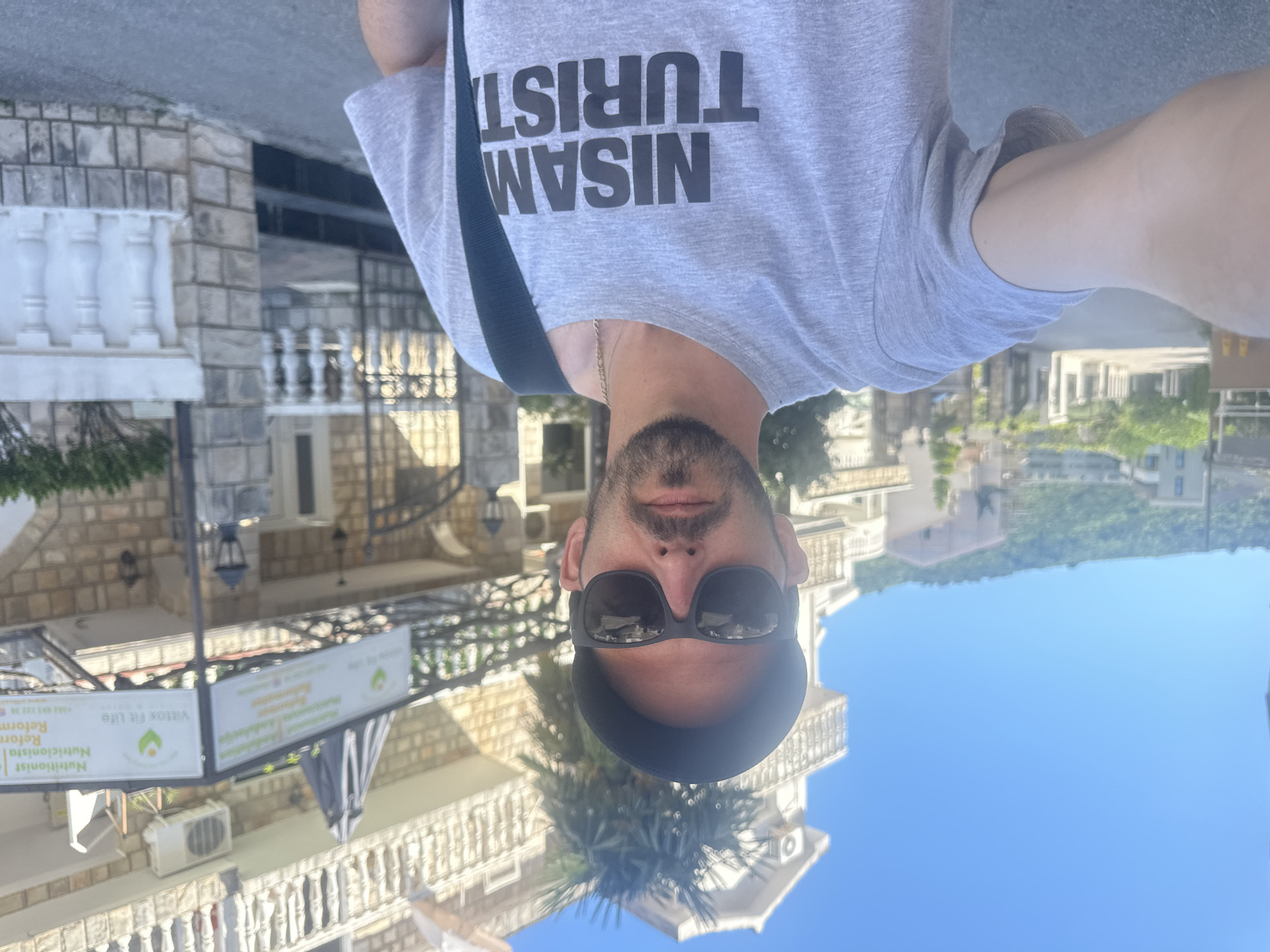 A man wearing sunglasses and a cap stands in front of a building in Budva, Montenegro. The man is wearing a gray T-shirt that reads "Nisam Turist" (I am not a tourist) in black letters. The building behind him has a sign that reads "Vitex Fit Life" in green letters. The sky is bright blue, and the sun is shining.