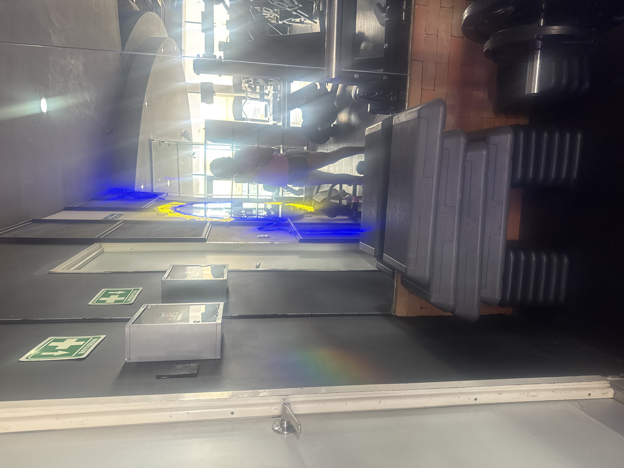 The image shows the interior of a gym in San Bernardino Tlaxcalancingo, Mexico. A woman exercises on a weight machine. Multiple weight plates and workout steps are visible. There are emergency exit signs on the wall. The background includes blue and yellow neon lights.