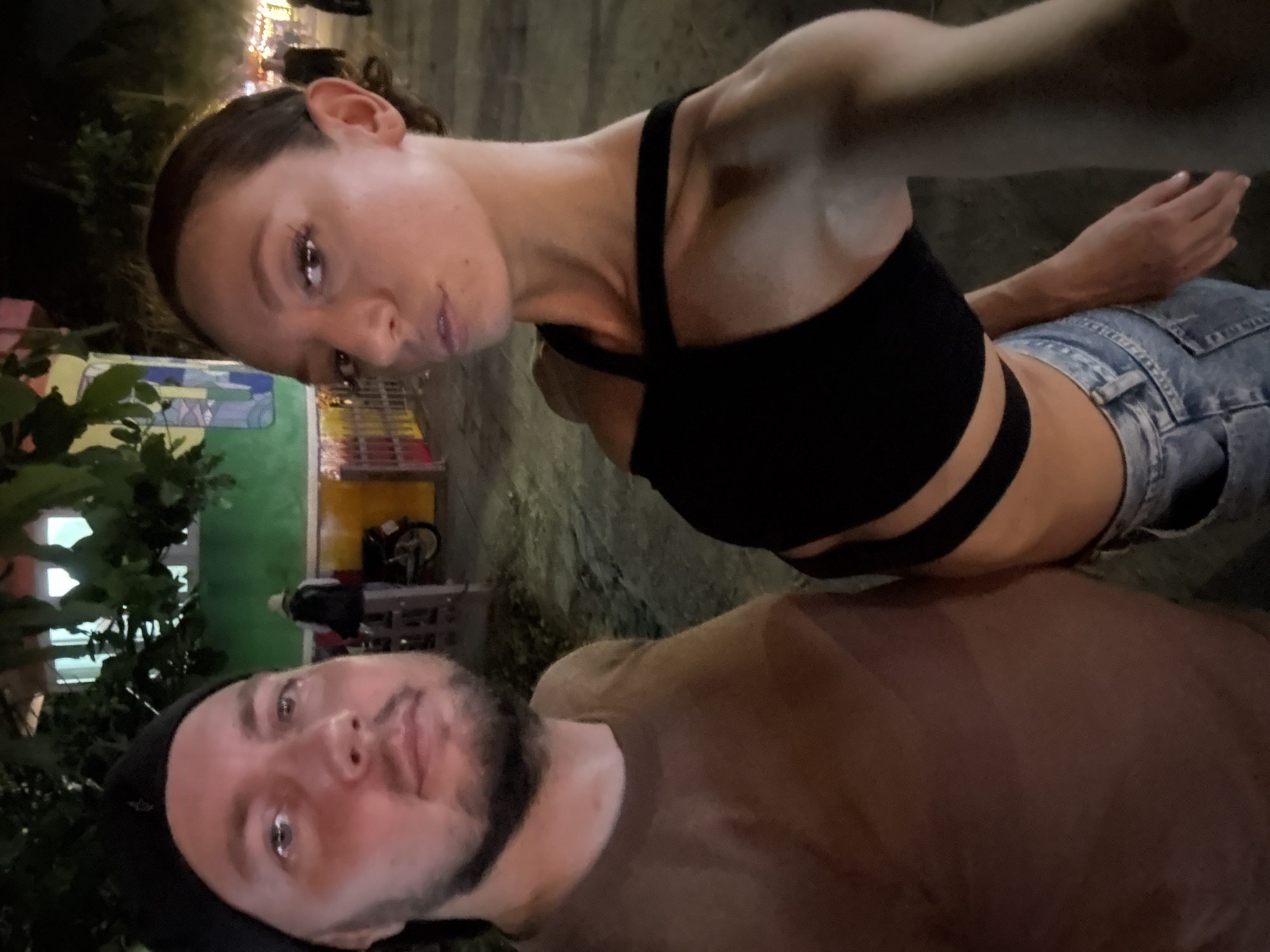 A selfie image taken outdoors at night shows a man and a woman in close proximity. The woman is in the upper portion of the frame, wearing a black crop top and denim shorts. The man, positioned in the lower portion, is wearing a brown t-shirt and a black cap. The background includes trees, buildings, and what appears to be street lighting. The image is rotated counterclockwise, making it appear sideways. The location is possibly in Đà Nẵng, Vietnam.