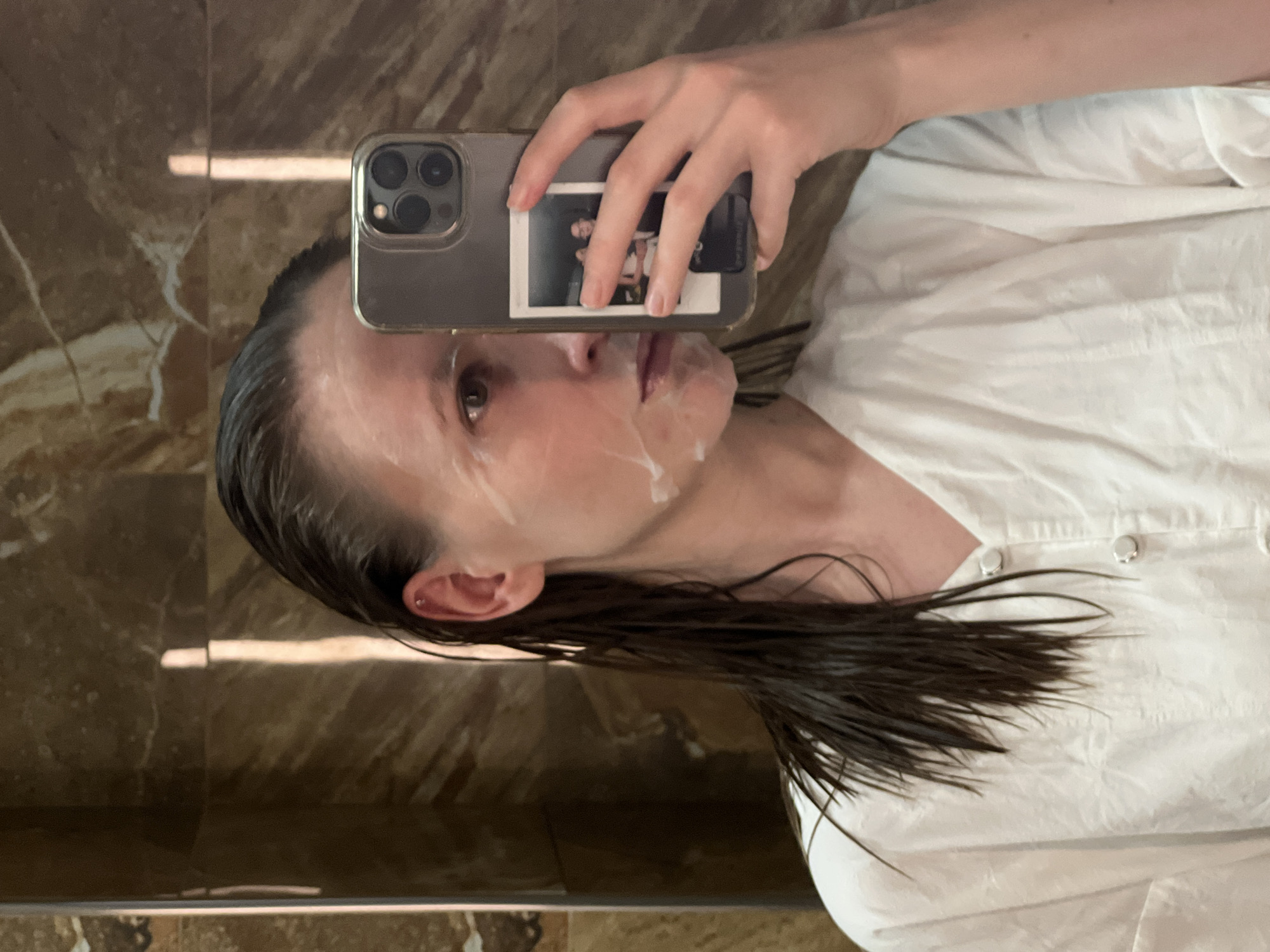 A person is taking a selfie in front of a mirror. The person has wet, dark hair and is wearing a white shirt. There is a white substance on the person's face, possibly a facial mask. The person is holding a smartphone with a photo attached to the back. The background appears to be a brown, marble-like surface, possibly a wall or countertop. There is no information that the location is in the city of Khimki, Russia.