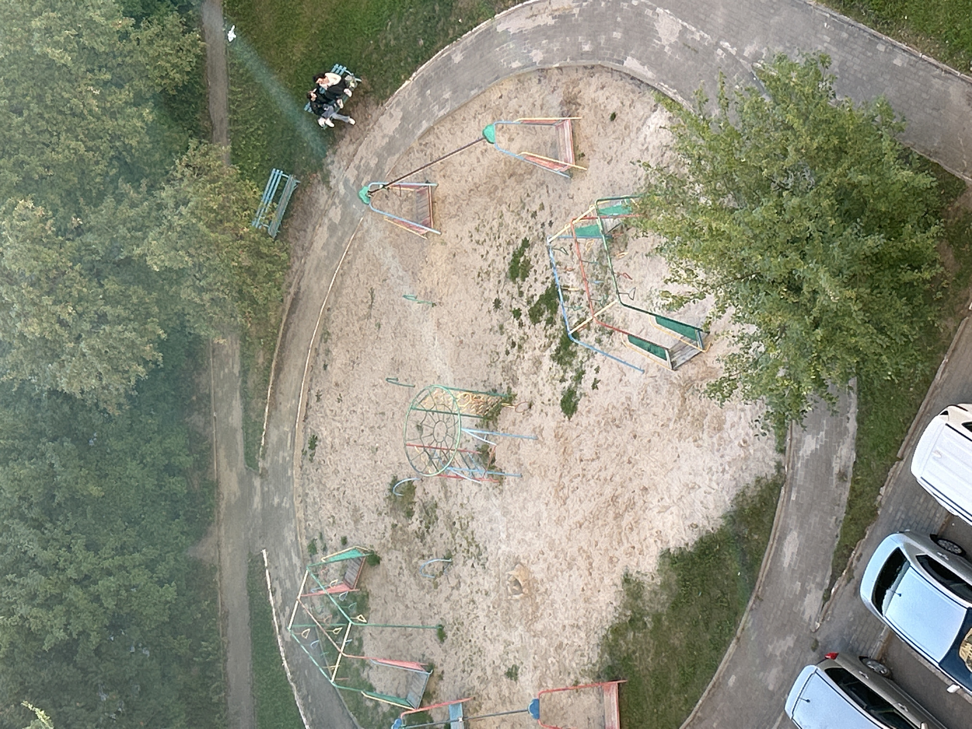 An aerial view captures a residential courtyard in Tomsk, Russia, featuring a playground and parked cars. The playground, situated in a sandpit, contains several swing sets, a climbing frame with a slide, and a climbing net structure. Two individuals are seated on a green bench on the grass near the playground, bathed in a shaft of sunlight. Several silver cars are parked along a paved walkway bordering the courtyard. Lush green trees and grass surround the area, indicating a natural, possibly summer or early autumn, environment.