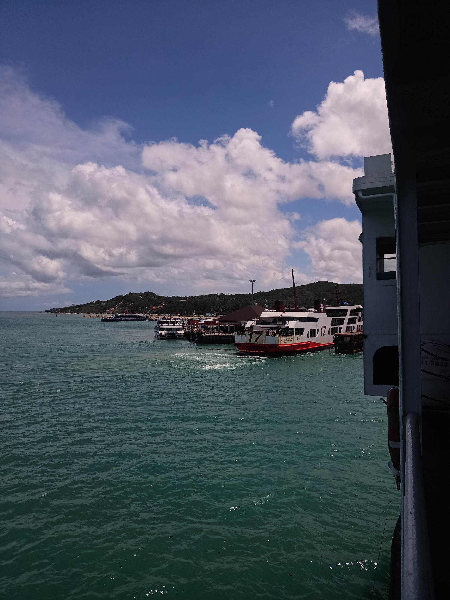 The image depicts a view from a ferry, likely docked or moving slowly near a pier in Thong Sala, Thailand. The turquoise water stretches out to the horizon, where a landmass with lush green vegetation is visible. Several ferries are docked at the pier, one prominently displaying the number "17" in large red lettering on its side. The sky above is filled with puffy white clouds against a blue backdrop. The side of the ferry from which the picture is taken is partially visible in the foreground, partially obscuring the scene.