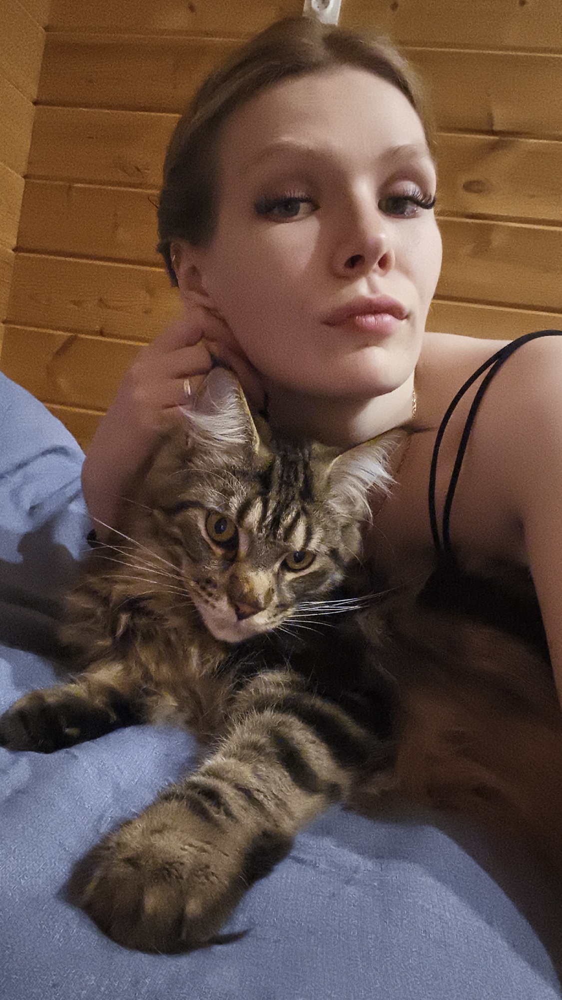 A woman with short brown hair, full lips, and heavy eyelash extensions is posing with a brown tabby Maine Coon cat. The woman is wearing a black tank top and has a gold necklace. She is lying on a blue surface, possibly a bed or couch, and her hand is near her face. The background consists of wooden planks. The location is unknown; Русский Вожой, Russia, cannot be confirmed from the image.