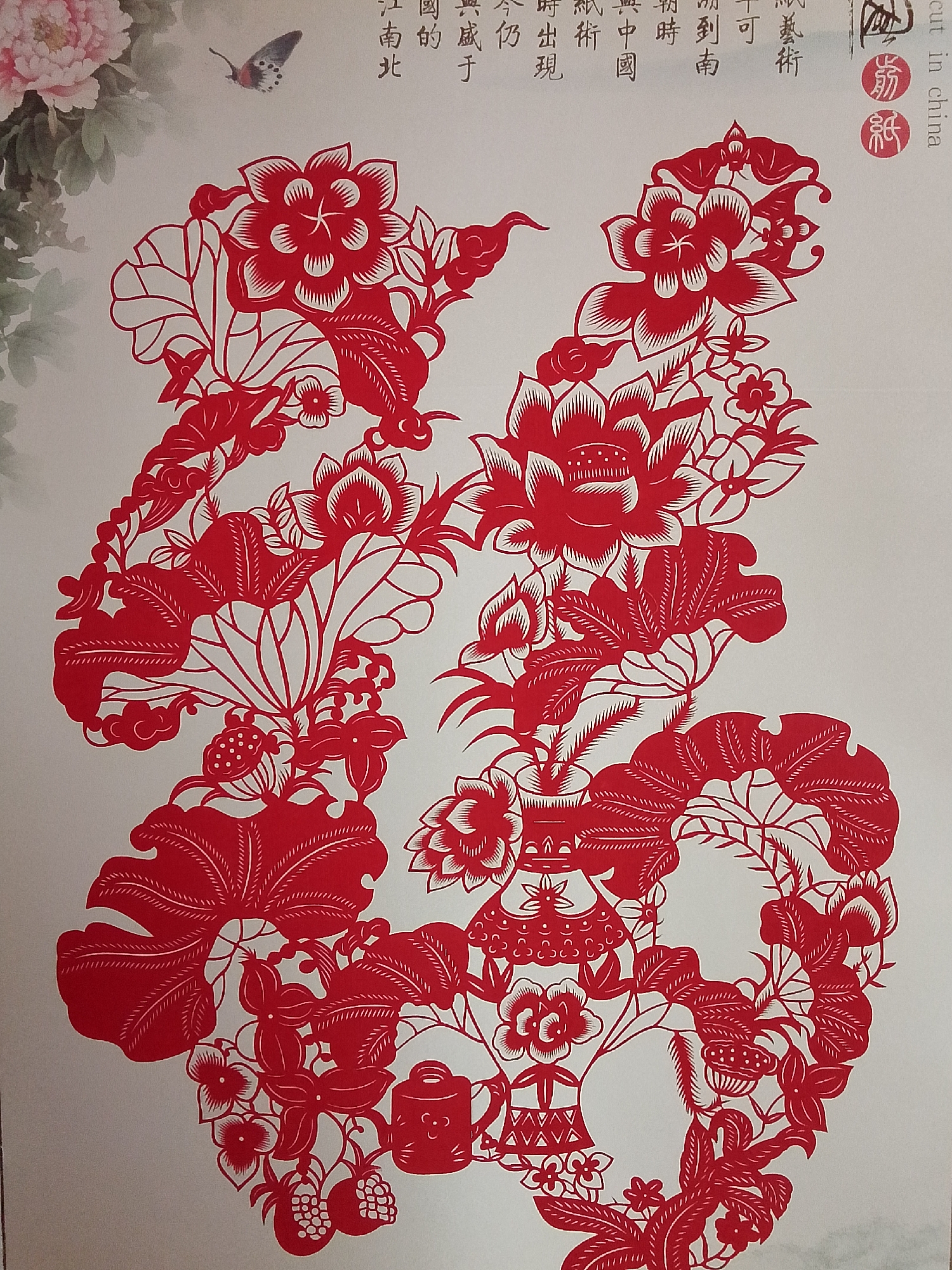 A detailed, red Chinese paper cut is displayed against a light background. The image includes floral motifs, leaves, and decorative elements arranged in a symmetrical design. There is also a butterfly in the upper left corner. The text on the top right side is Chinese and "cut in china" written in English.