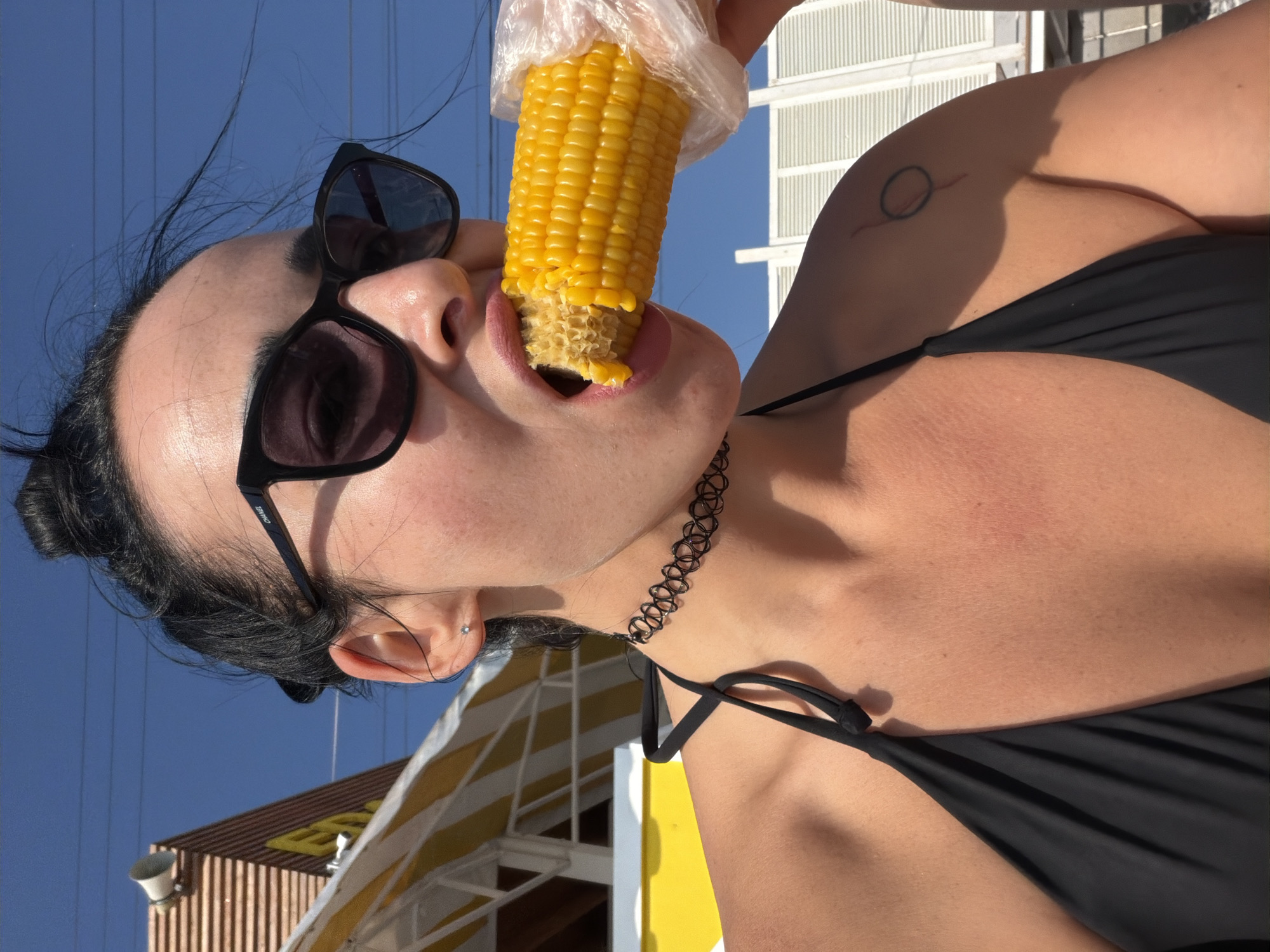 A woman with dark hair in a bun, wearing sunglasses, a black choker necklace, and a black bikini top, eats corn on the cob. She is outdoors under a clear blue sky, with what appears to be a beachside establishment with yellow and white striped awnings in the background. The scene is likely in Sochi, Russia, given the context.