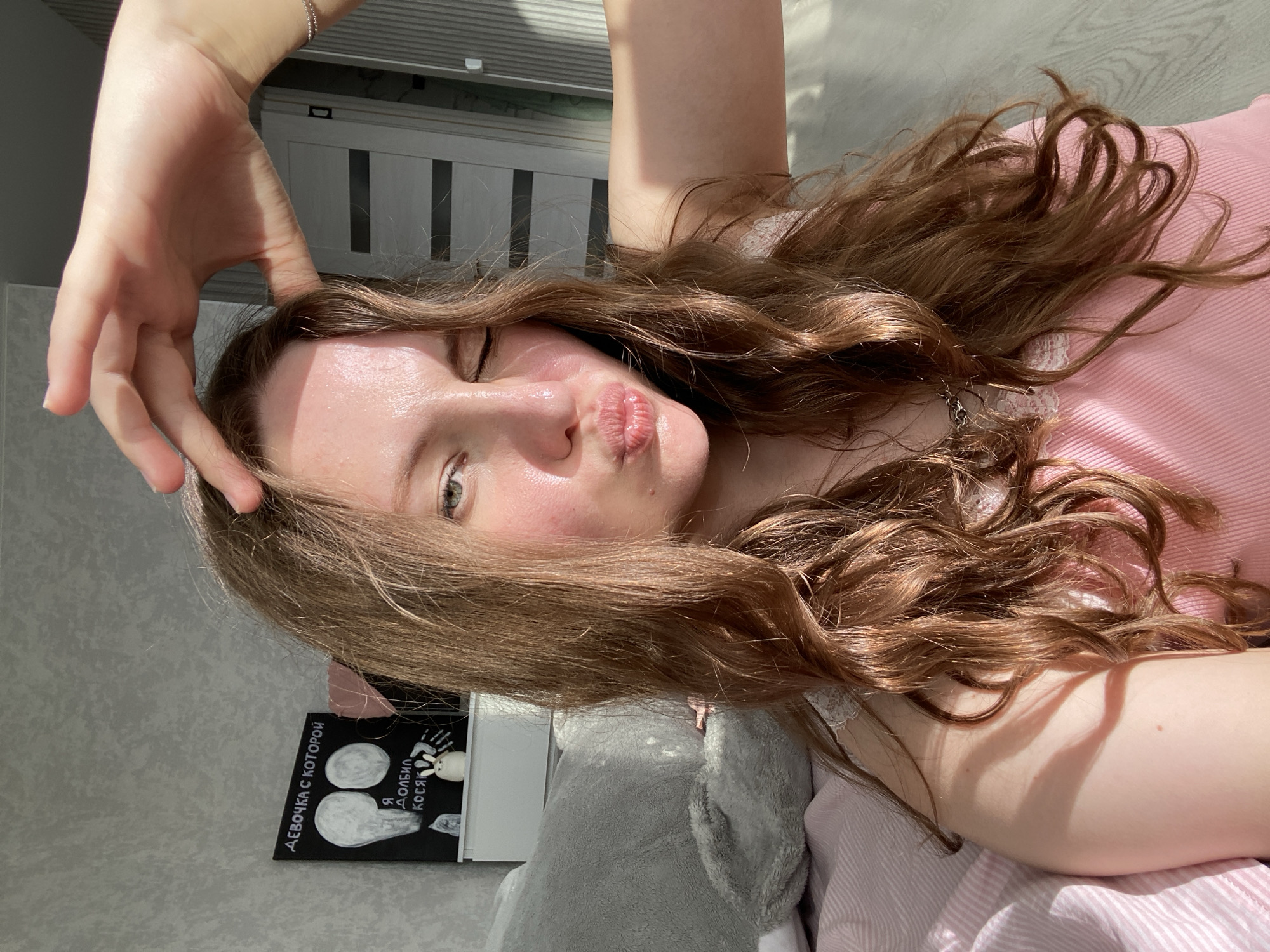 The media file is a selfie taken by a young woman with long, wavy, brown hair. She is lying down, with her head tilted back, and her right arm raised, touching her forehead. Her eyes are closed, and her lips are pursed, giving her a playful expression. She is wearing a light pink top with lace trim.
The background appears to be a bedroom. There is a grey stuffed animal visible, and a pink and white striped blanket or sheet. To the left, a dark framed object with white drawings and Russian text is partially visible. The text appears to be "ДЕВОЧКА С КОТОРОЙ" and "Я ДОЛБИЛ КОСЯК". The lighting suggests it is daytime, with sunlight casting shadows across the scene. The overall mood is relaxed and casual.
The context of Izhevsk, Russia, provides a geographical location for the image.
