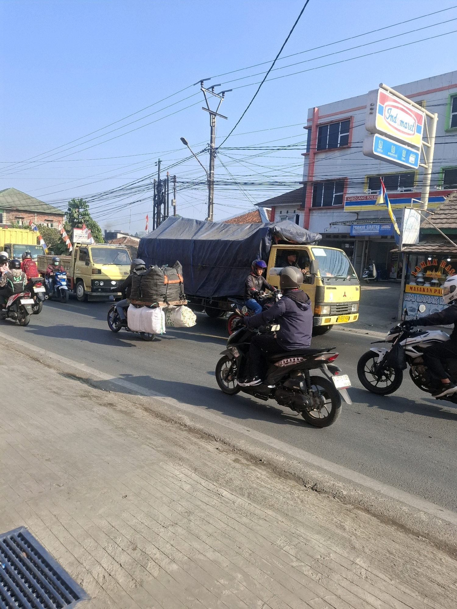 The image captures a street scene in Selajambe, Indonesia, characterized by moderate traffic under a clear blue sky. In the foreground, a gray concrete sidewalk with a metal grate is visible on the left. The road itself is asphalt, with a clear lane marking suggesting multiple lanes of traffic.
Several motorcycles are present, with riders wearing helmets. One motorcycle in the mid-ground is carrying a significant load of bundled goods, appearing to be sacks. Another motorcycle in the foreground is black and has the word "STREET" visible on its side. A white and black motorcycle is to the right.
A yellow truck is positioned in the center of the frame, with its cargo covered by a dark tarp. A man is visible driving the truck. A large, dark tarp-covered truck is behind the yellow truck, suggesting a convoy or heavy transport.
On the right side of the road, buildings line the street. One building features a prominent "Indomaret" sign, indicating a convenience store. Another building appears to be a shop with signage that is partially visible and in Indonesian. Flags are also visible, including Indonesian flags. Power lines and utility poles are scattered across the scene, common in urban and semi-urban environments. The overall atmosphere is that of a typical day with ongoing commerce and transportation.