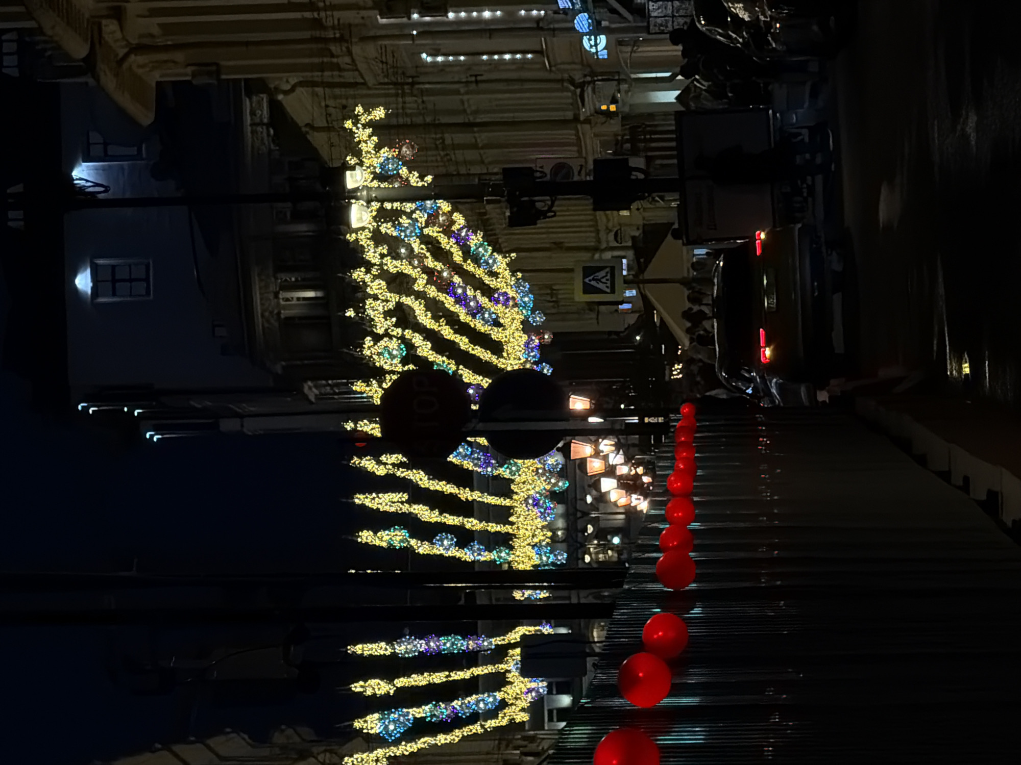 The image captures a night scene in Moscow, Russia, likely during a holiday season due to the festive decorations. A brightly illuminated Christmas tree-like structure adorned with yellow and blue lights dominates the view. The tree is centrally located on a street lined with buildings, one of which has lit windows. Further down the street, there's a row of red spherical decorations. The street seems busy with cars, some of which are visible with their headlights and tail lights on. Pedestrians are also present. The lighting suggests it is night.