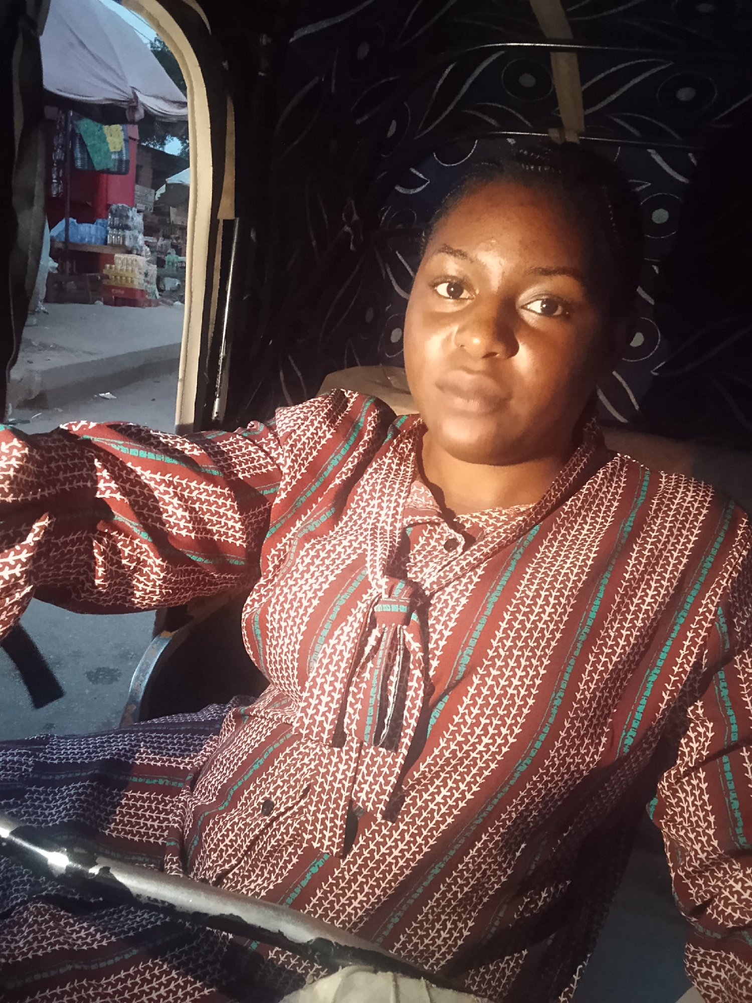 A woman is seated inside a vehicle, possibly a motorized tricycle, with a partial view of a bustling commercial area outside. The woman is wearing a patterned shirt with burgundy, white, and turquoise colors and a tied neckline. Outside the vehicle, there are visible commercial stalls with goods displayed, indicating a marketplace. The scene appears to be in Bukuru, Nigeria, based on contextual knowledge of typical local transportation and commercial environments. The lighting inside the vehicle is subdued compared to the brighter daylight outside.