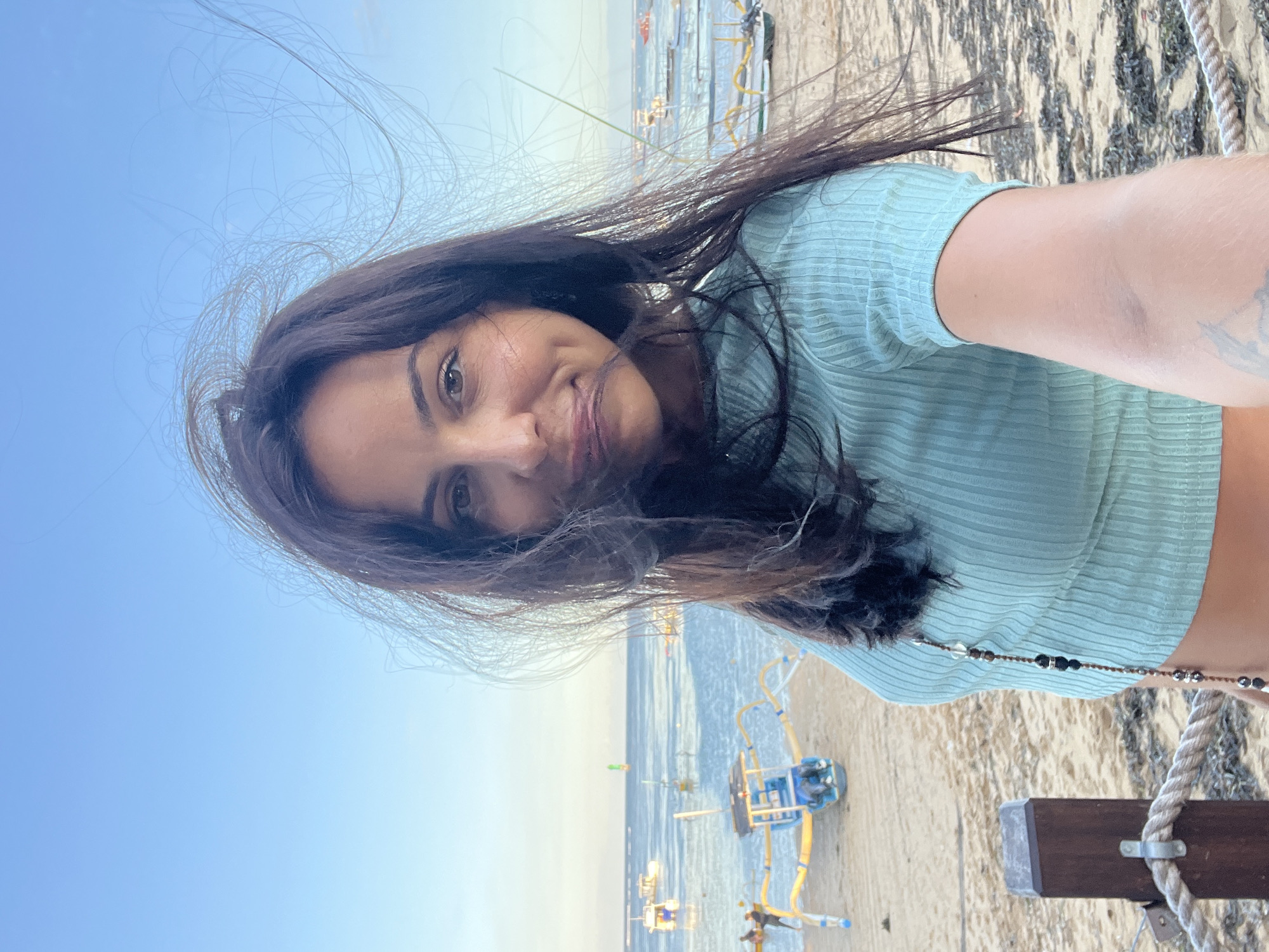 A woman is standing on a beach in Denpasar, Indonesia, taking a selfie. She has dark hair that is being blown in the wind, and she is wearing a light blue cropped top. Behind her is a sandy beach with several small boats in the water. There is a wooden post with a rope railing near the bottom right of the frame. The sky is light blue.