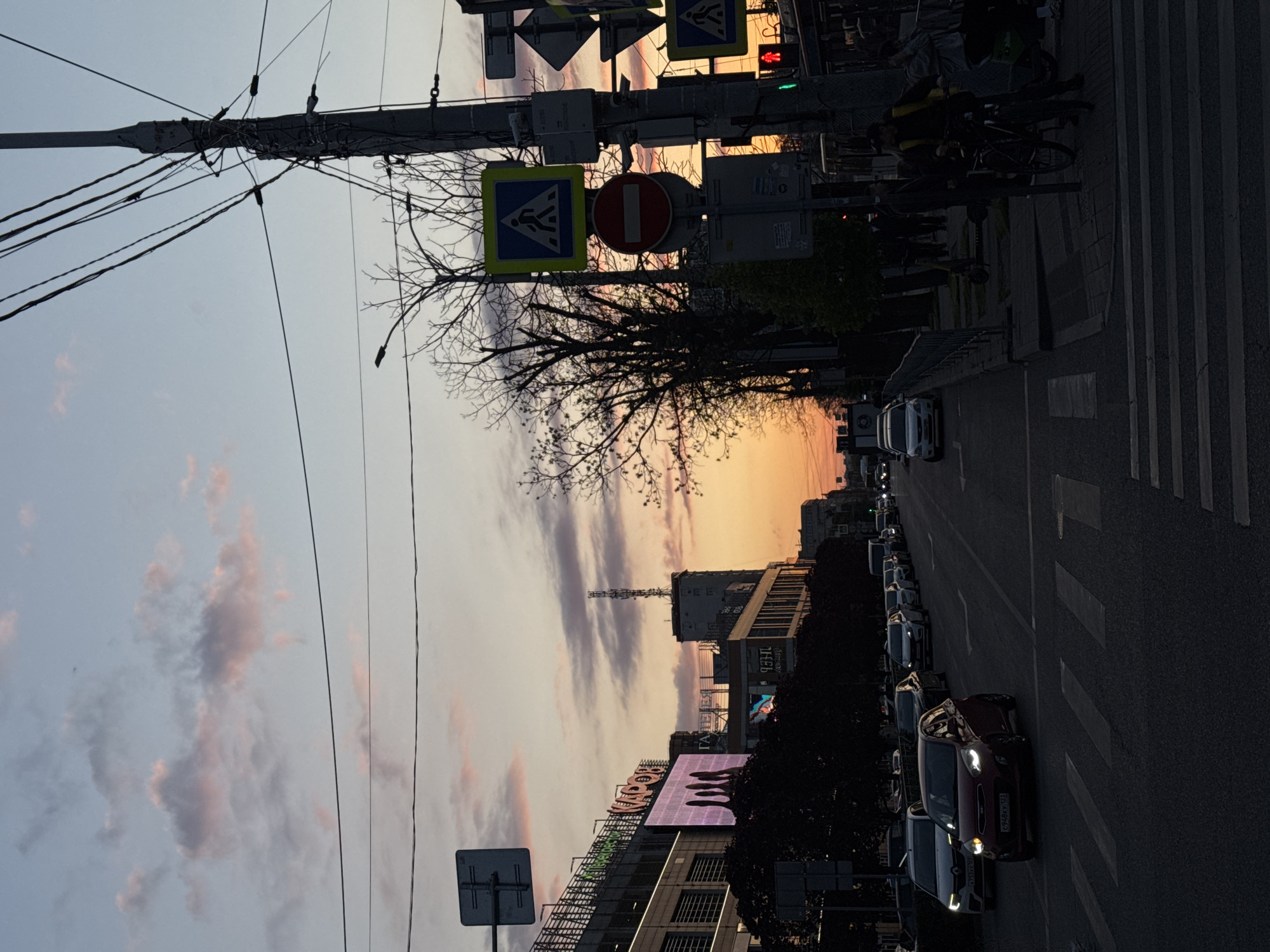 The image depicts an urban street scene during sunset or dusk, characterized by the warm, transitioning light. The sky exhibits a gradient of colors, with vibrant orange and pink hues near the horizon fading into lighter blues and scattered greyish-pink clouds overhead.
In the foreground, a utility pole heavily laden with numerous electrical wires extends across the upper left, while a bare tree with intricate branches is silhouetted against the colorful sky. Several traffic signs are mounted on poles, including a blue and yellow pedestrian crossing sign, a red "no entry" sign, and a traffic light displaying a red pedestrian signal.
Modern buildings line the street, some featuring illuminated screens and visible signage. Text like "ЦЕНТР ЦИФРОВОГО ПАРК" (Center of Digital Park) and "ТЕХН" (Tech) is discernible on one building facade, and another sign partially shows "ЯНДЕКС". A cellular tower stands on the roof of a building in the mid-ground.
The asphalt road below is marked with white crosswalk stripes. Various cars are parked along the curb, and several vehicles are driving, including a prominent red car in the middle ground with its headlights on. Its license plate is partially visible as "С948ОР 762". On the sidewalk, indistinct figures of people can be seen, some appearing to be on bicycles or scooters, indicating a typical evening commute. The scene conveys a bustling city transitioning from day to night.
