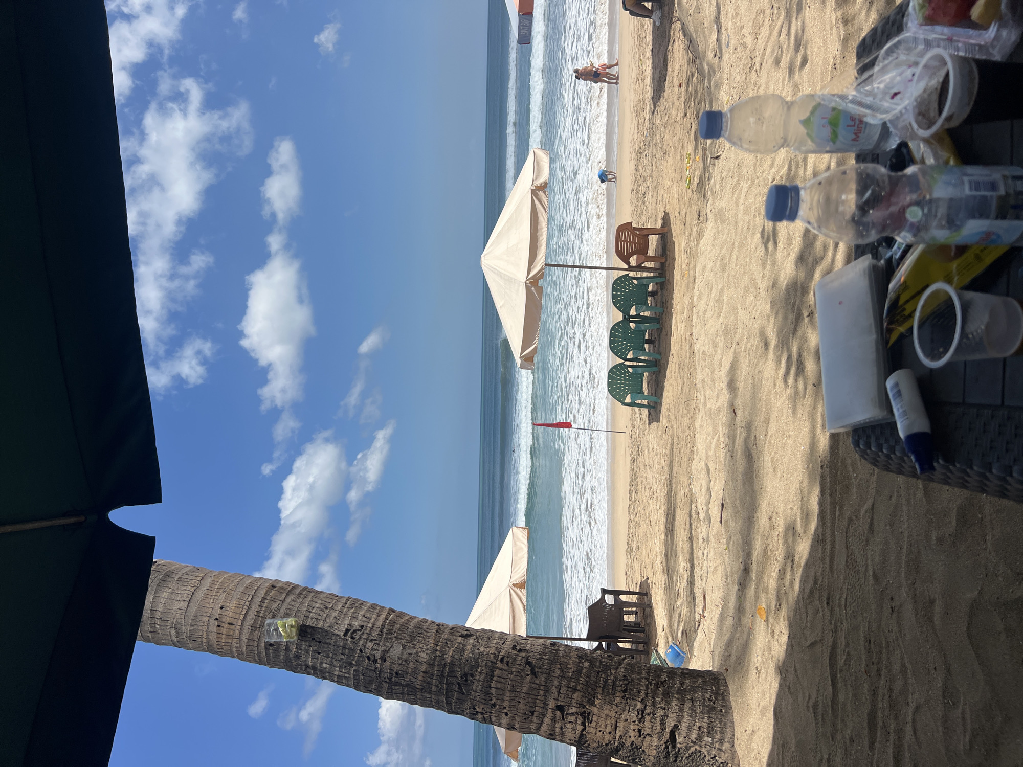 The image depicts a sunny day on Kuta Beach, Bali, Indonesia. In the foreground, a woven black table holds two plastic bottles of water, one partially filled, and two disposable cups, one with a dark beverage. A white packet and a yellow bag are also on the table. A large palm tree trunk stands on the left, its rough bark textured and casting a shadow. A small clear cup with green pieces, possibly fruit or ice, is attached to the trunk. The sandy beach stretches towards the ocean, where gentle waves are breaking. Several beach chairs, three green and one brown, are arranged near a beige beach umbrella. A red flag stands planted in the sand. In the distance, people can be seen on the beach and in the water, including a child playing near the shoreline and two adults walking. The sky is a vibrant blue with scattered white clouds. The overall atmosphere suggests a relaxed day at the beach.