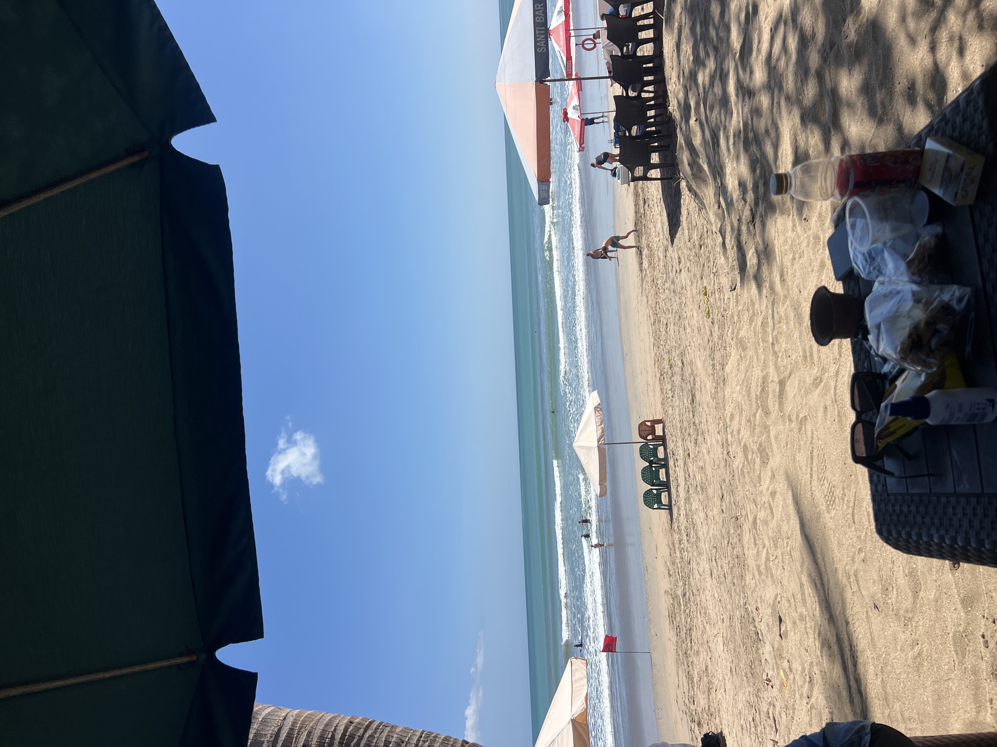 The image depicts a beachfront scene in Kuta, Indonesia. In the foreground, a dark rattan table holds various items including sunglasses, a bottle of red liquid, a plastic cup, a small potted plant, a phone, and two plastic bags containing food items. Further back, the sandy beach is dotted with several beach umbrellas. To the left, a tall palm tree is partially visible. In the middle ground, there are rows of plastic chairs, a red flag planted in the sand, and the ocean with gentle waves. Several individuals are visible on the beach and in the water, some wading or swimming, and others walking or playing. The sky is clear blue with a single white cloud. The scene suggests a leisurely day at the beach.
