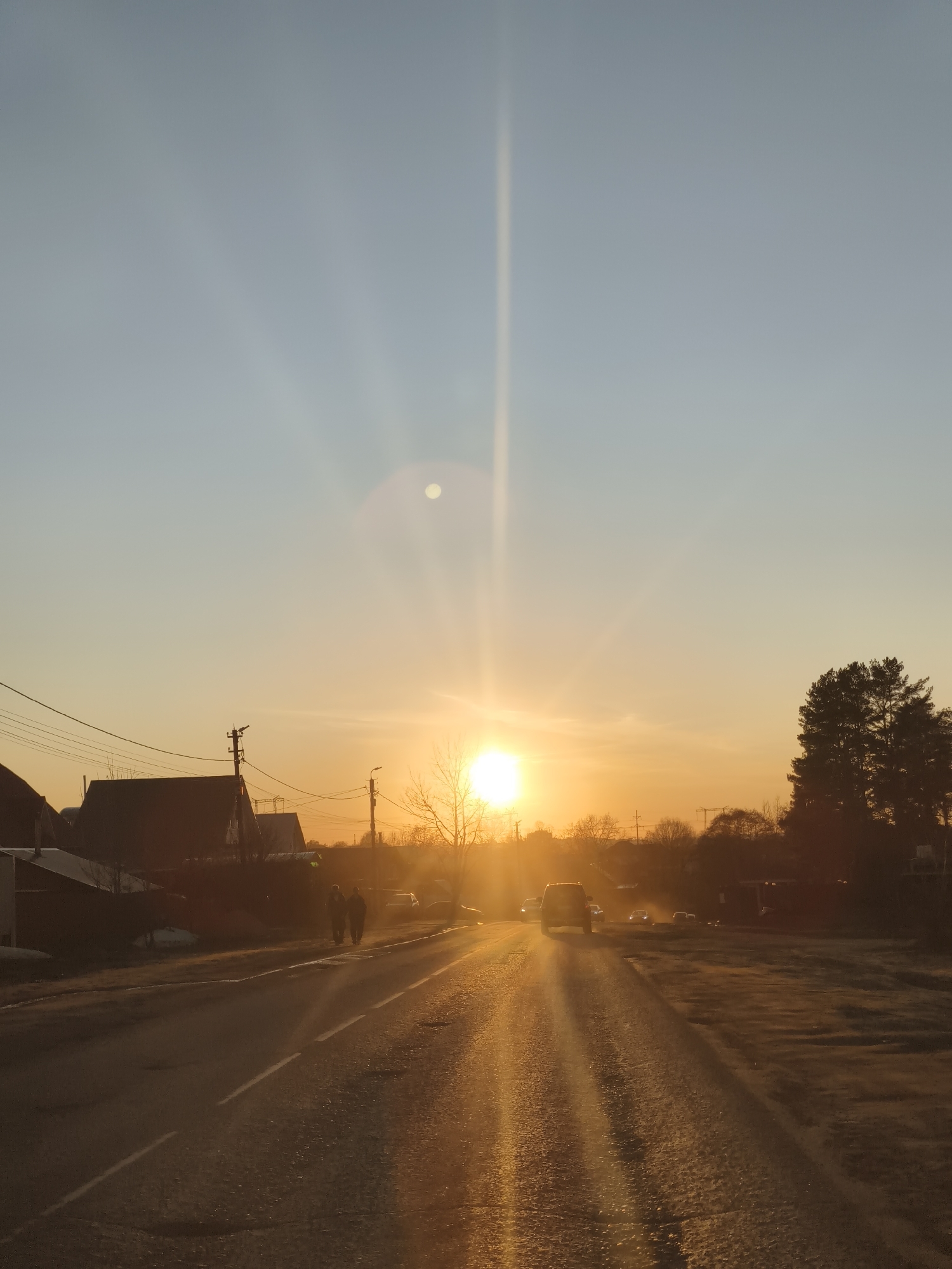 The image depicts a road in a town, likely at sunrise or sunset, based on the warm golden sunlight and long lens flare extending vertically from the sun. The sky transitions from light blue to orange near the horizon. Silhouetted houses and trees line the left and right sides of the road, respectively. Two individuals are walking on the left side of the road, with several cars traveling in both directions. A car is approaching the camera from the front. The location appears to be Unknown, Unknown based on the lack of recognizable landmarks.