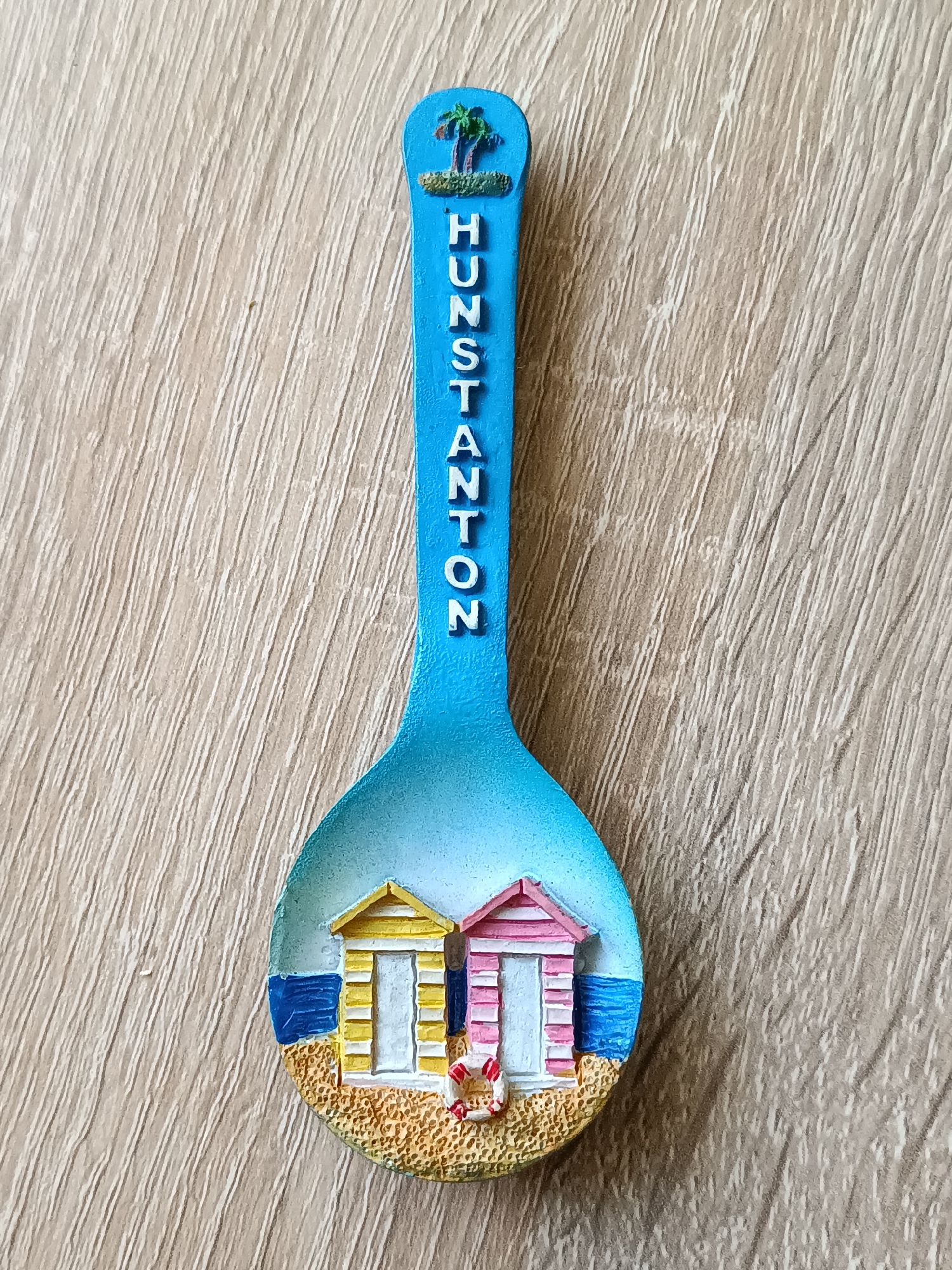 A souvenir spoon with a depiction of Hunstanton beach huts and the text "HUNSTANTON" on its handle rests on a light brown wooden surface. The spoon is a bright blue color and features a small palm tree at the top of its handle. The bowl of the spoon displays two colorful beach huts, one yellow and one pink, situated on a sandy beach with a blue sea in the background. The scene is a static, decorative representation of a coastal location. The wooden surface exhibits a grain pattern. The lighting is from above, casting subtle shadows.