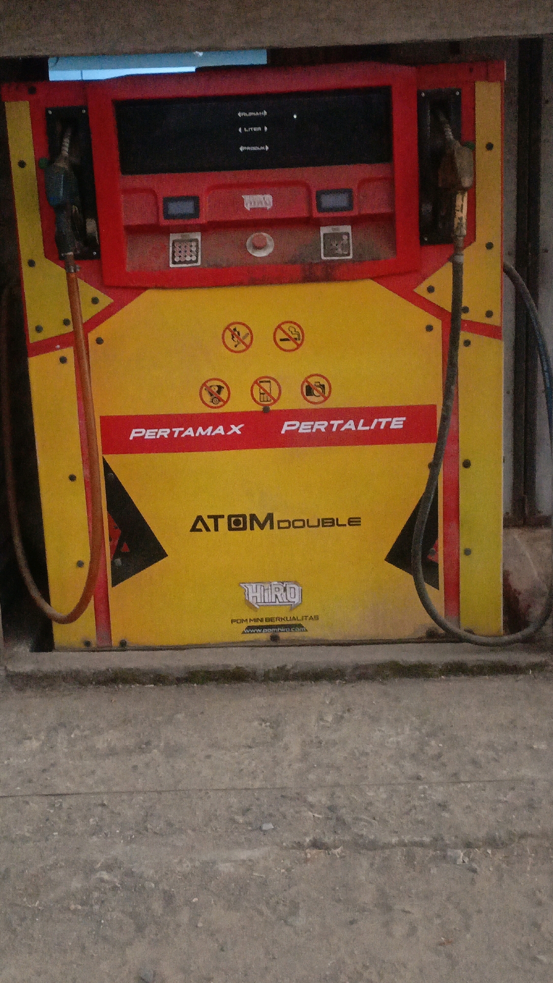 The image depicts a yellow and red Pertamina gas pump, a common sight at fuel stations in Indonesia, including the city of Taliwang. The pump is an upright, double-sided unit with separate nozzles for Pertamax and Pertalite fuel. The Pertamax side is on the left and the Pertalite side is on the right. The pump displays information about fuel prices and volumes on a digital screen. Safety icons are visible on the yellow panel, including "No Smoking" and "No Open Flames." The gas pump is situated outdoors, likely at a roadside fuel station, with a concrete ground surface in the foreground and a dark, possibly brick or concrete wall in the background. The overall impression is of a functional and well-maintained piece of public infrastructure.