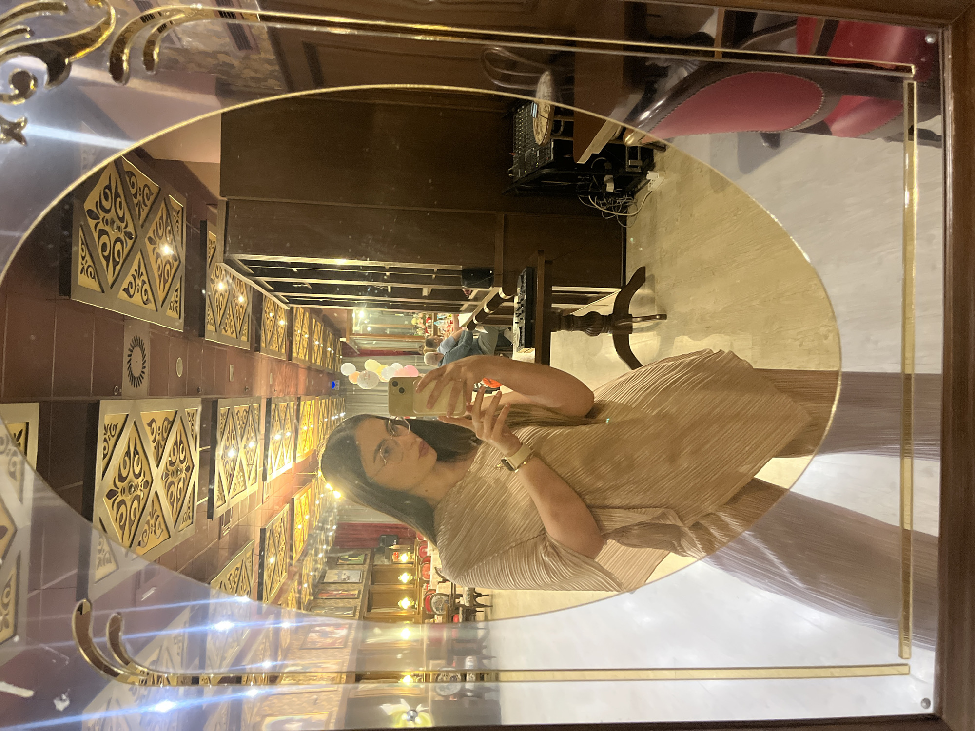 A mirror reflects a woman in a beige pleated dress taking a selfie with her smartphone. The woman wears glasses and a wristwatch. The reflection reveals an elaborately decorated interior, possibly a restaurant or hotel lobby, with a long hallway featuring panels with gold and dark brown designs. A few individuals are visible in the background of the reflection, along with balloon decorations. The surroundings are well-lit, and the mirror itself has a decorative gold frame. The location is in the city of Unknown, Unknown.