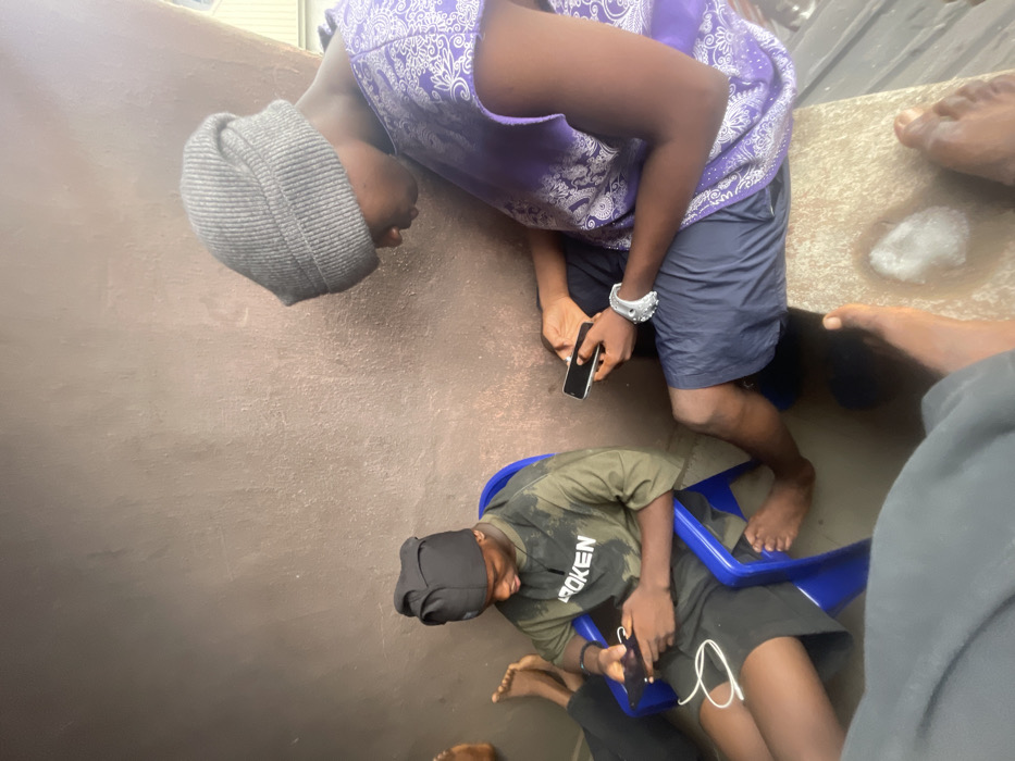Two individuals are seen from above, occupied with their phones. One wears a gray beanie, a purple top, and navy shorts, while the other sits in a blue plastic chair, sporting a black head covering, an army green t-shirt, and black shorts. Both are intensely focused on their devices. Additional bare feet and lower body parts of other people are partially visible around them, suggesting a crowded space.