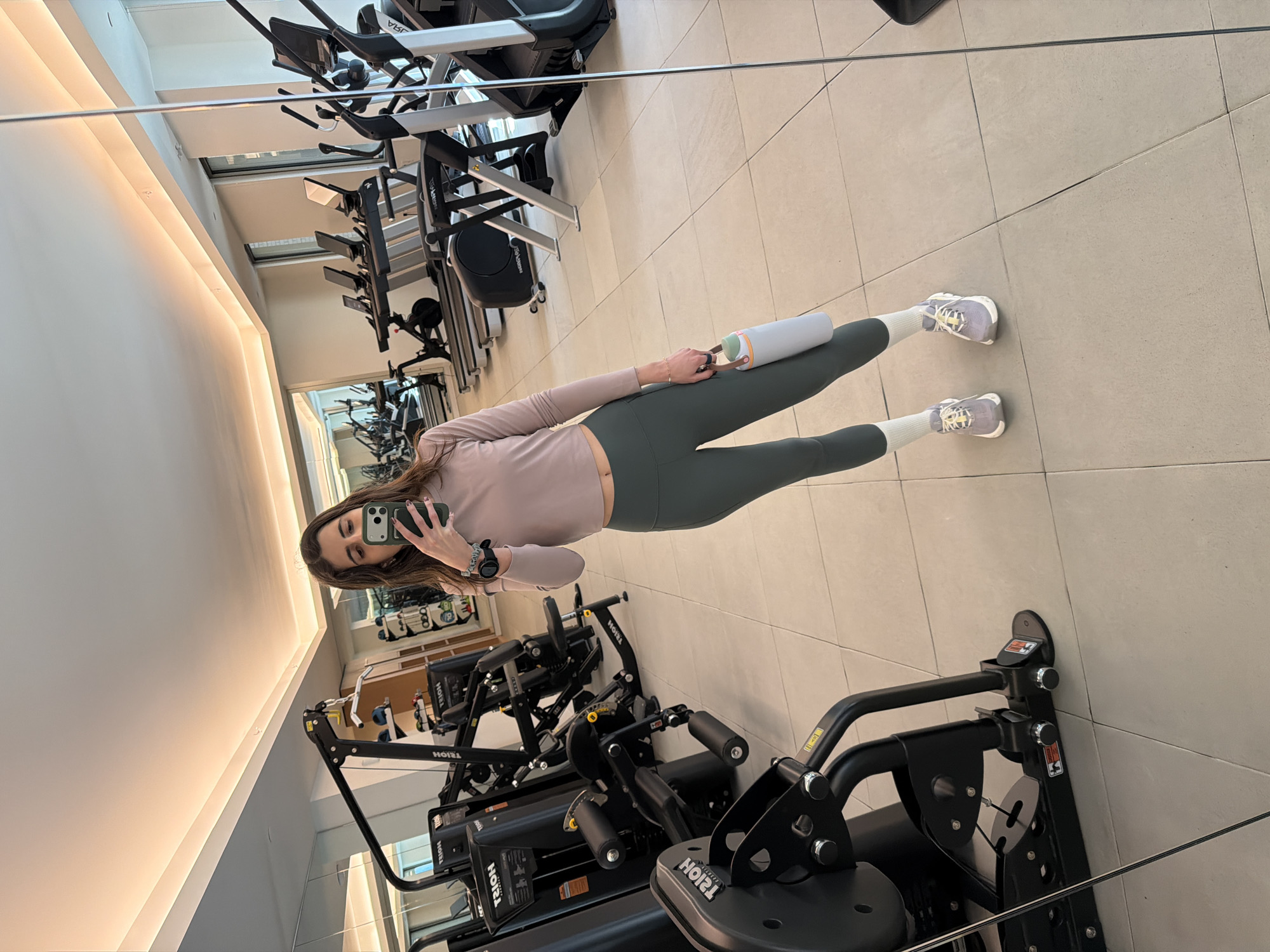 A woman is taking a selfie in a gym. The gym has various exercise machines, including treadmills and weight machines. The floor is tiled. The woman is wearing a long-sleeved shirt, leggings, socks, and sneakers. She is holding a water bottle. The location is most likely in New York City, United States of America, due to the architectural style commonly seen in the city's gyms and fitness facilities.