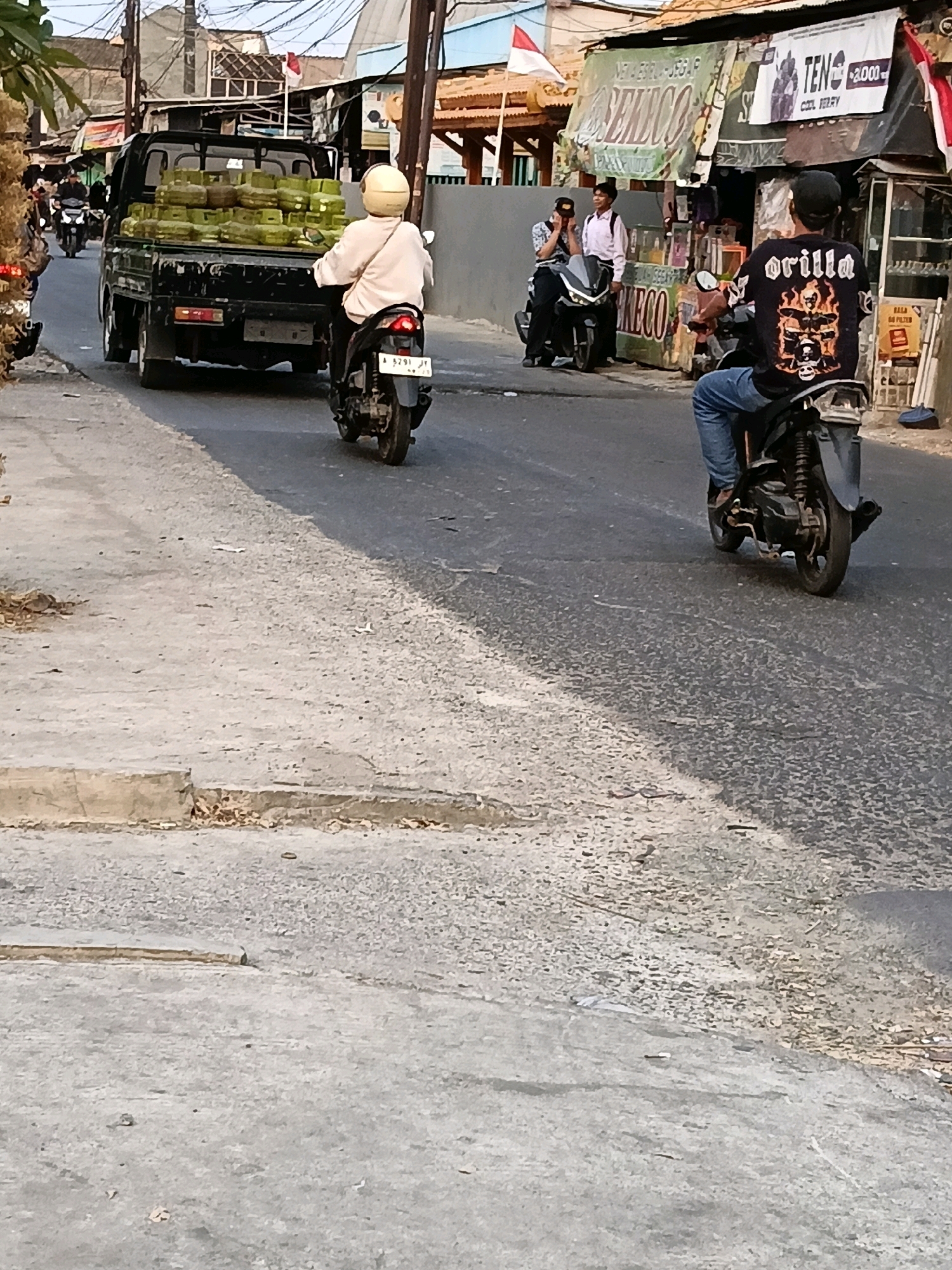 A black utility truck loaded with green propane tanks drives down a paved street in Belimbing, Indonesia. A person wearing a light-colored shirt and a yellow helmet rides a motorcycle behind the truck. Another person wearing a black t-shirt with an "Orilla" graphic and blue jeans rides a motorcycle in the opposite direction. The buildings lining the street appear to be small shops or residences. The weather is bright, suggesting daylight.