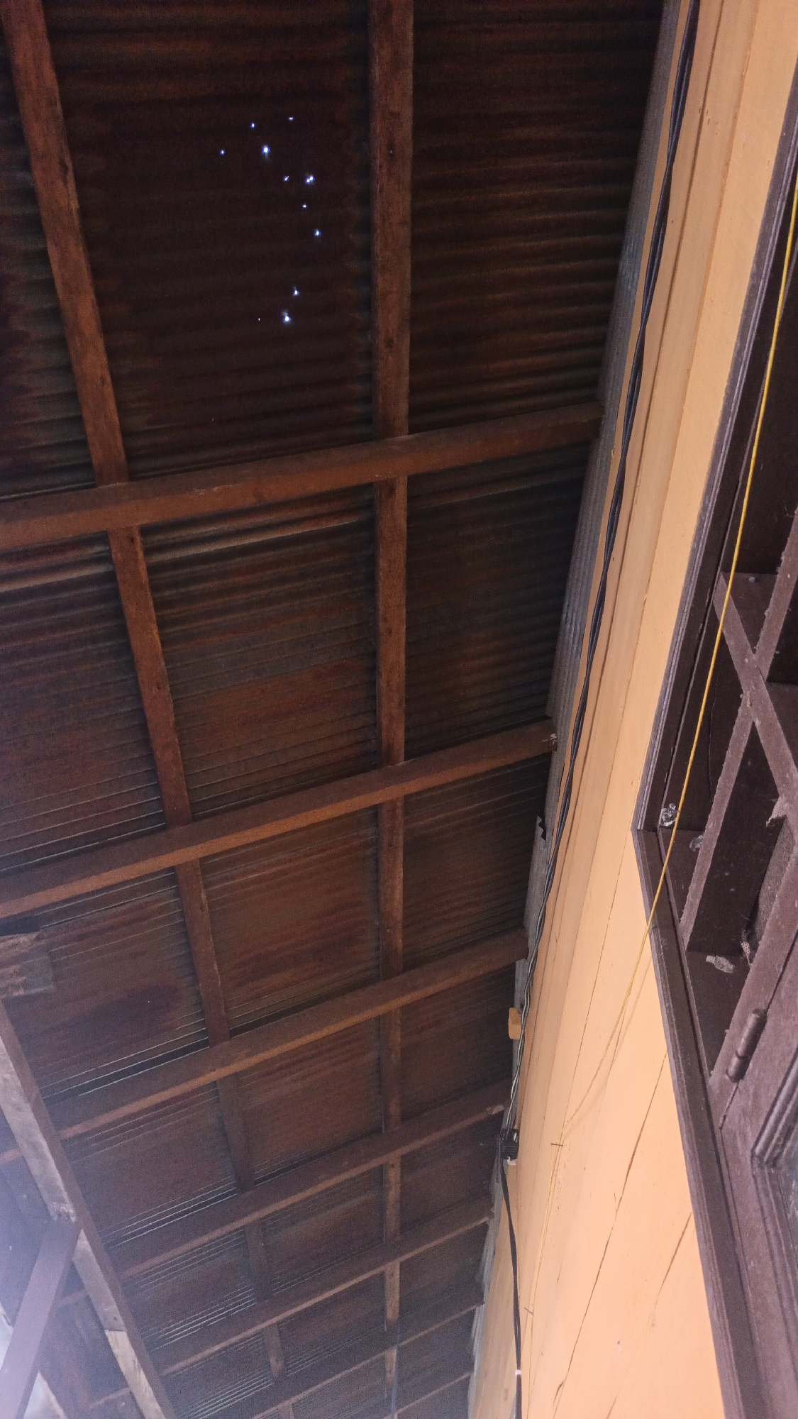 The image shows an under view of a weathered corrugated metal roof supported by wooden beams. The roof is dark brown and shows signs of age and rust. Several bright, star-like points of light are visible on the underside of the roof in the upper left portion of the image, potentially from a light source or insects. To the right, a section of an orange-painted building wall is visible, with some electrical wiring attached. A dark brown window frame with a lattice design is also present on the right side of the image. The scene suggests an outdoor, possibly covered, area. The presence of an orange building and potentially common building materials in Banjarmasin, Indonesia, suggests a location within the city.