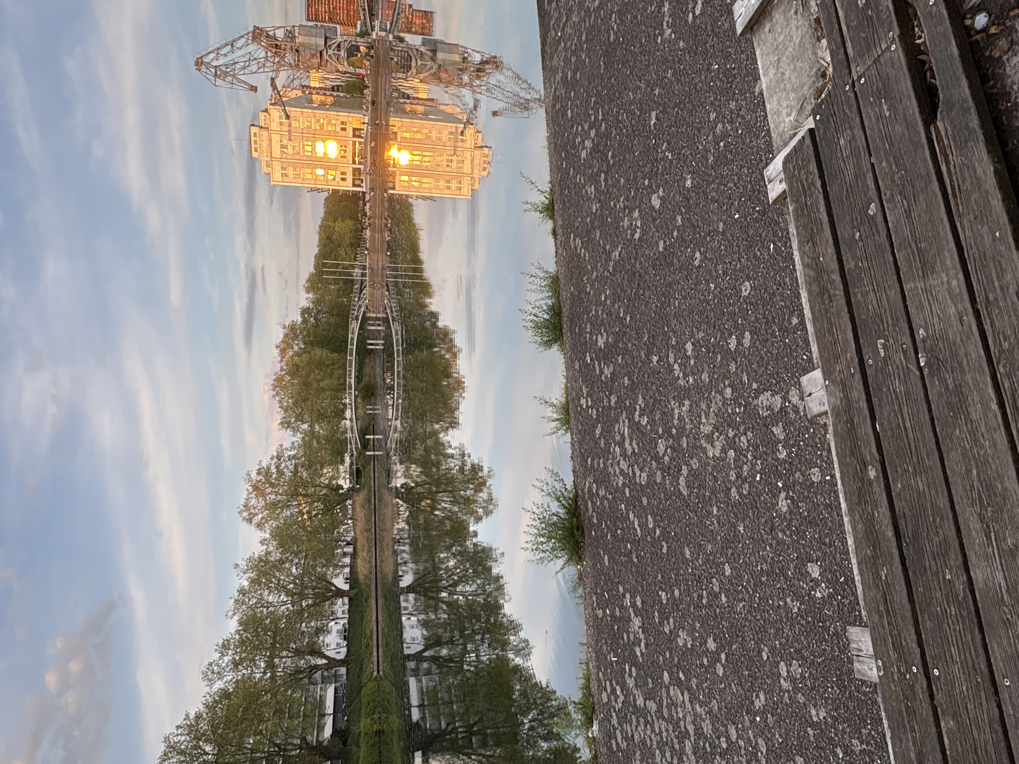 The image shows a reflection of a scene in Strasbourg, France, likely in a body of water. Reflected in the water is a building with windows illuminated, a large crane structure, trees lining a path, and the sky with clouds. The actual ground, not the reflection, is visible in the lower right, composed of a dark asphalt surface and a wooden bench. The image appears to be taken from a low angle near the ground.