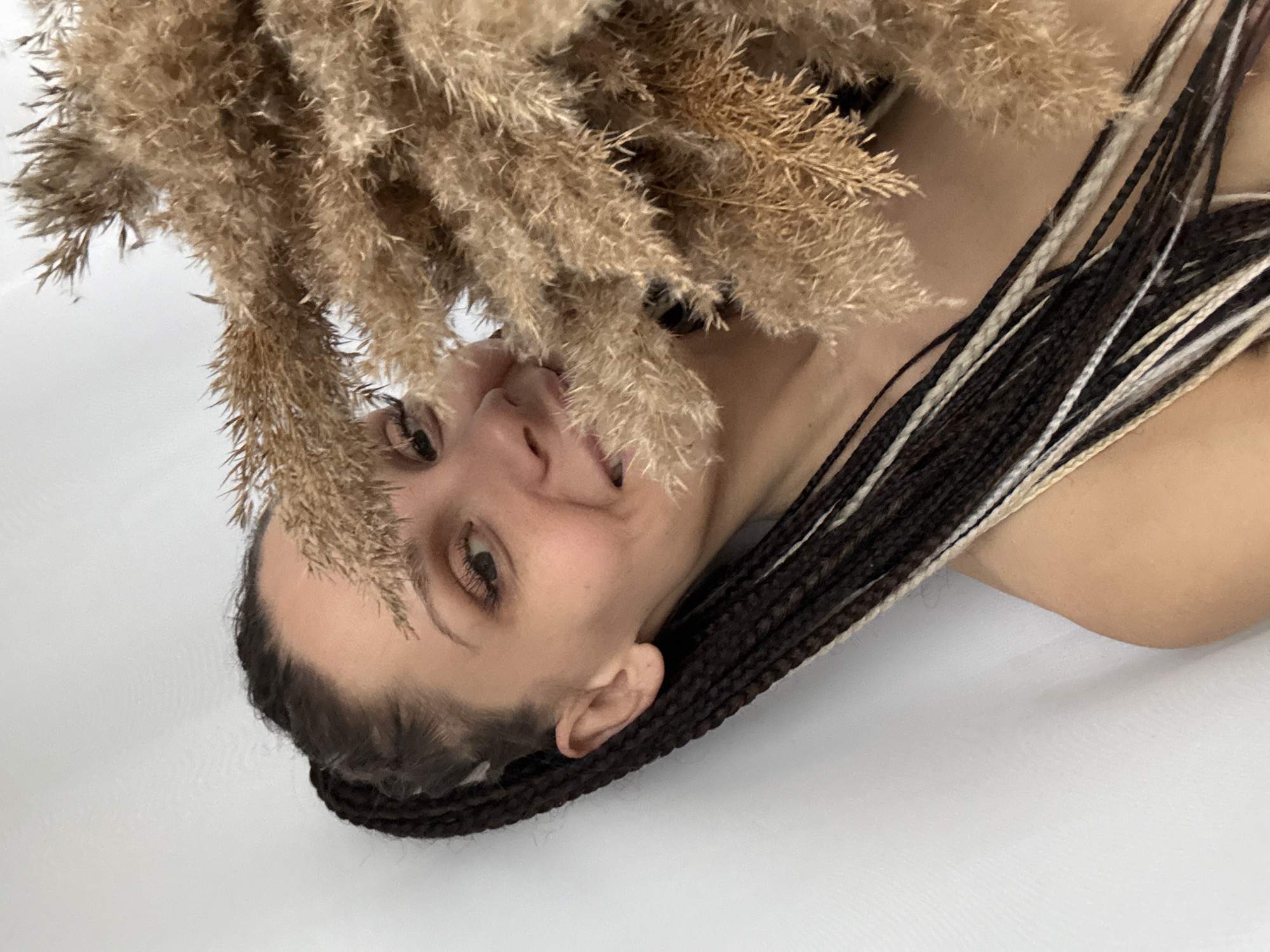 A woman with dark and light braided hair is positioned in front of a white background. A bunch of dried grass-like foliage partially covers her face. The location is unknown.