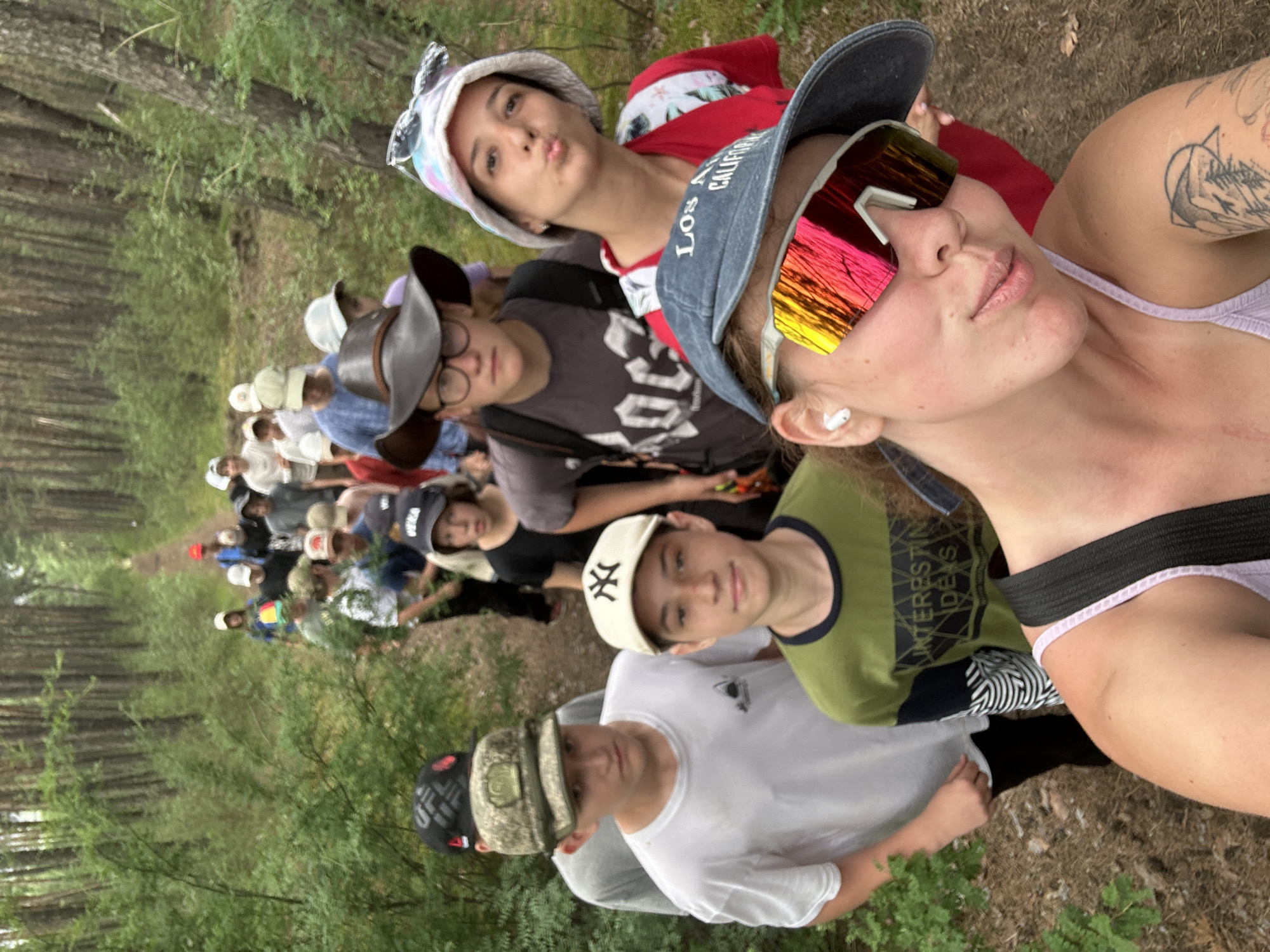 In a forest setting, a group of young people walks in single file on a dirt path. The foreground is dominated by the close-up of a young woman wearing a blue cap and large, colorful sunglasses. Her mouth is pursed, as if making a kissy face, and she is looking at the camera. Her left shoulder is visible, showing a tattoo of a mountain scene. Behind her, a young person wearing a green shirt with "INTERESTING IDEAS" printed on it looks forward. To the left of them, another young person in a white t-shirt also faces forward. Further up the path, a line of individuals, mostly teenagers and young adults, are visible, some wearing hats and backpacks. The environment is densely wooded with tall pine trees and undergrowth on either side of the path. The lighting suggests it is daytime, with dappled sunlight filtering through the trees. The location is identified as being in the city of Nikolo-Petrovka, Russia.