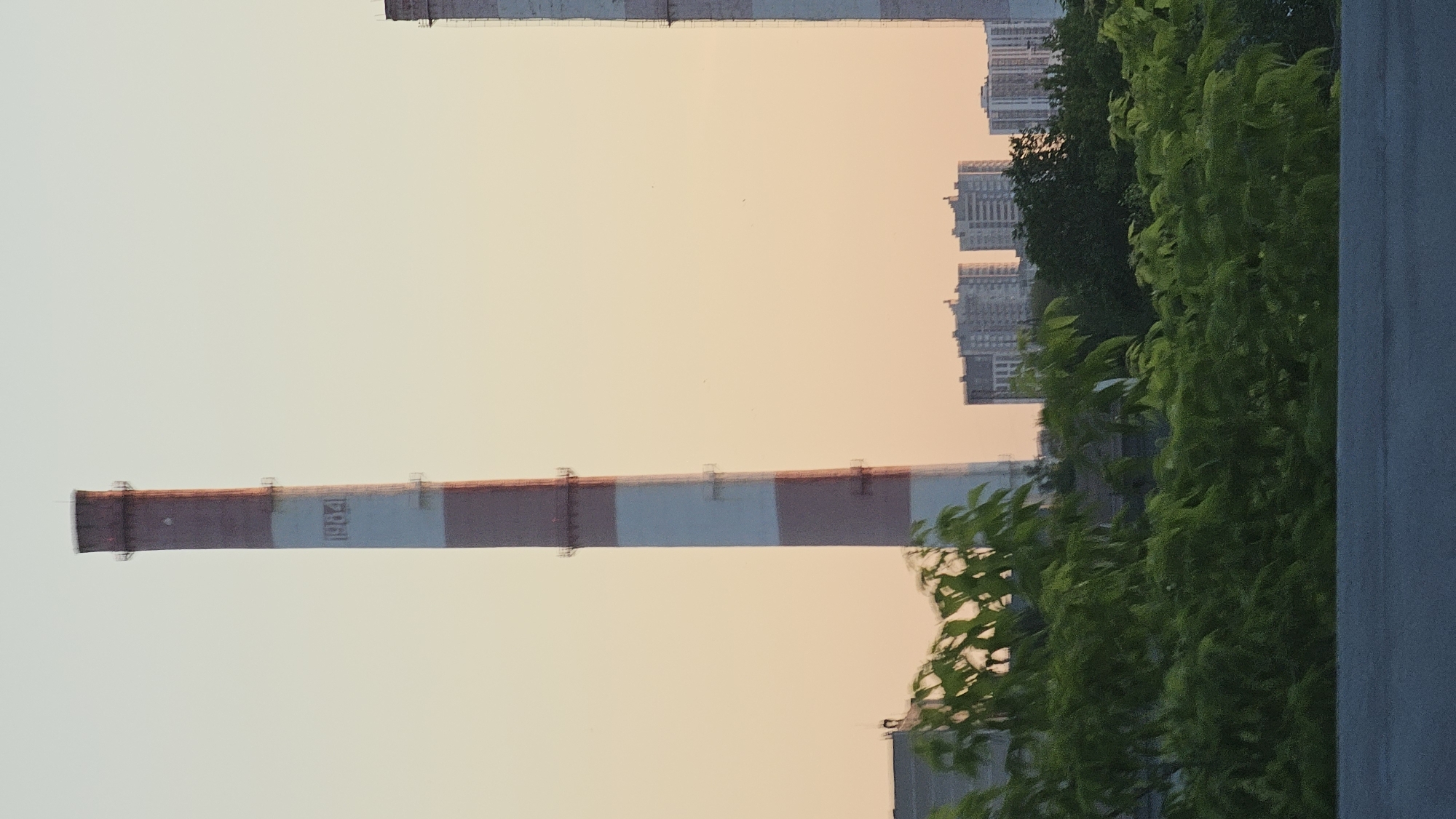 The image depicts a portion of the Yekaterinburg CHPP-2 thermal power plant, a prominent landmark in Yekaterinburg, Russia. In the foreground, lush green foliage obscures the base of the scene, with a dark gray paved surface in the very bottom. Dominating the upper left quadrant is a tall, red and white striped smokestack, bearing the year "1984" in dark lettering on one of its white bands. To the right, another similar smokestack is partially visible, also appearing red and white. In the mid-ground, behind the foliage, are several multi-story buildings, likely residential or commercial structures, with distinct architectural styles. The sky is a pale, soft orange, indicative of either sunrise or sunset. There are no people or visible actions in the image. The overall impression is one of industrial architecture set against a backdrop of urban development and natural elements.