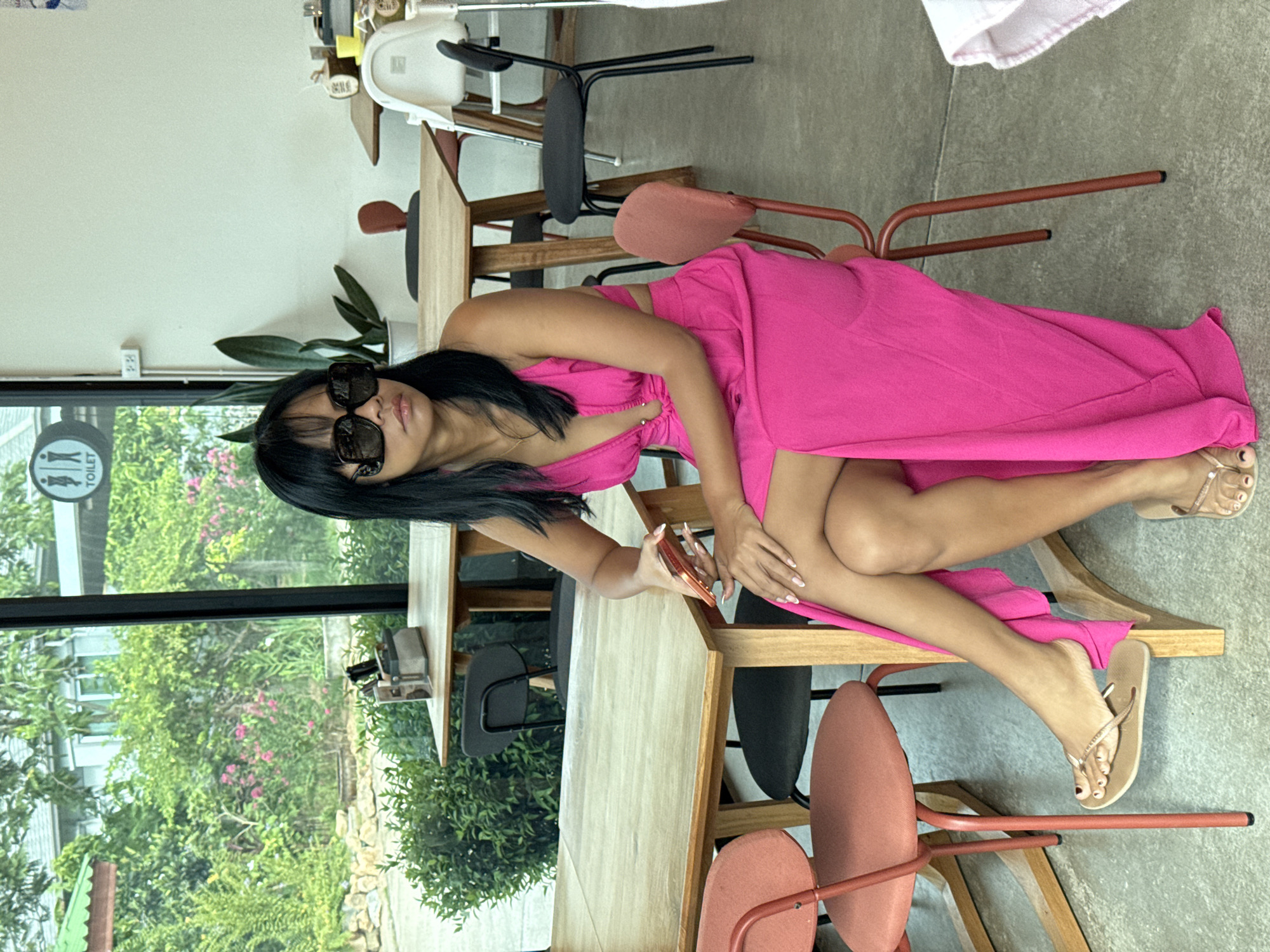 A woman with dark hair and sunglasses sits at a wooden table in a cafe, wearing a bright pink dress and flip-flops. She holds a mobile phone in her right hand. The cafe appears to be located in Ko Samui District, Thailand, with a view of lush greenery through a large window. The interior features modern furniture, including wooden tables and chairs with red accents. The floor is concrete.