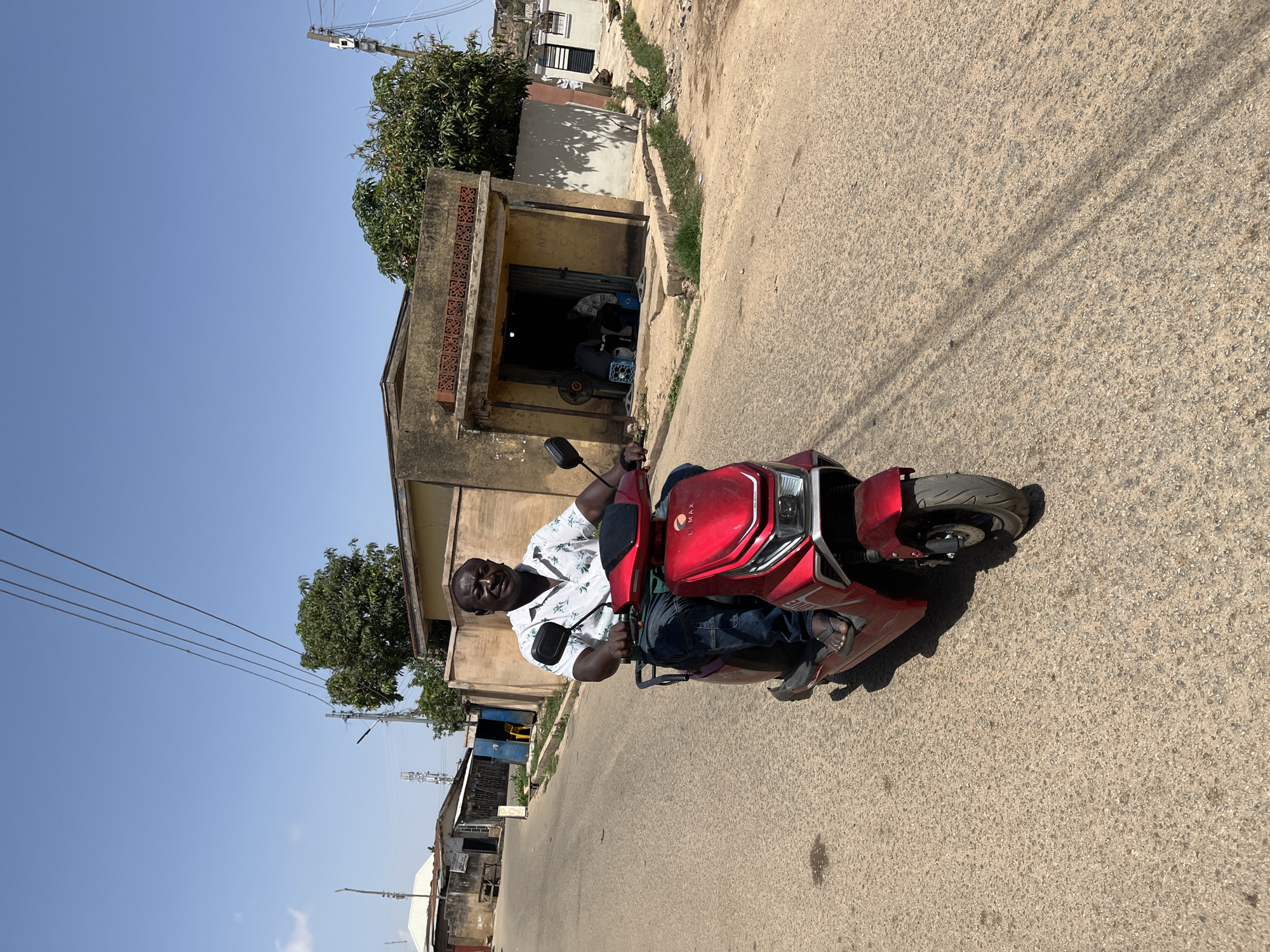 A medium shot depicts an adult man riding a red LMAX motorcycle on a paved road. He wears a patterned white short-sleeved shirt and blue jeans, along with dark-colored sandals. The man is facing forward, looking directly at the camera. The setting appears to be a street in Jos, Nigeria. Buildings line the side of the road, including a yellow building with a dark opening, possibly a shop or workshop. Power lines are visible overhead. The sky is clear and blue. The road shows signs of wear.