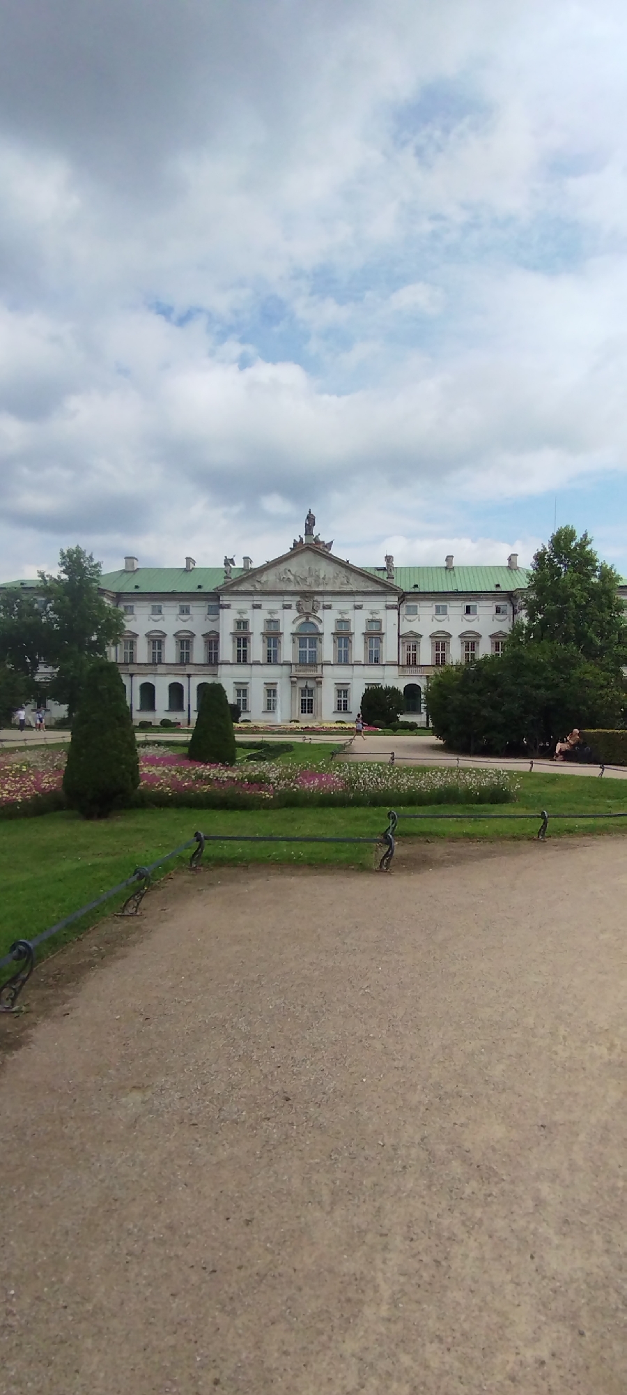 This image shows a view of the Royal Castle in Warsaw, Poland, on a cloudy day. The castle is a large, white building with a green roof, featuring classical architecture with numerous windows and a central pediment decorated with sculptures. In front of the castle, there is a meticulously manicured garden with flower beds and trimmed conical trees. A gravel pathway runs through the foreground, bordered by a decorative metal fence. Several people are visible in the scene, some walking in the garden and one couple sitting on a bench. The sky is filled with dramatic grey and white clouds, suggesting overcast weather.